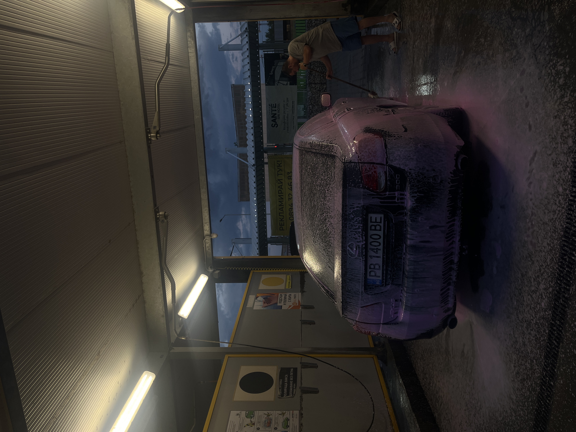 The image depicts the back of a dark-colored car, covered in foam and seemingly undergoing a car wash, inside a car wash bay. A man is partially visible in the background, appearing to be using a long-handled brush to wash the car. The license plate number is PB 1400 BE. The bay is lit by fluorescent lights. Several advertisements and instructions on a panel in the bay are for a "VODOSRUJKA" system. The background includes an advertisement for "SANTE SPA HOTEL" in Velingrad, Bulgaria, suggesting the car wash is located near Plovdiv, Bulgaria. The image suggests the interior of a self-service car wash facility.