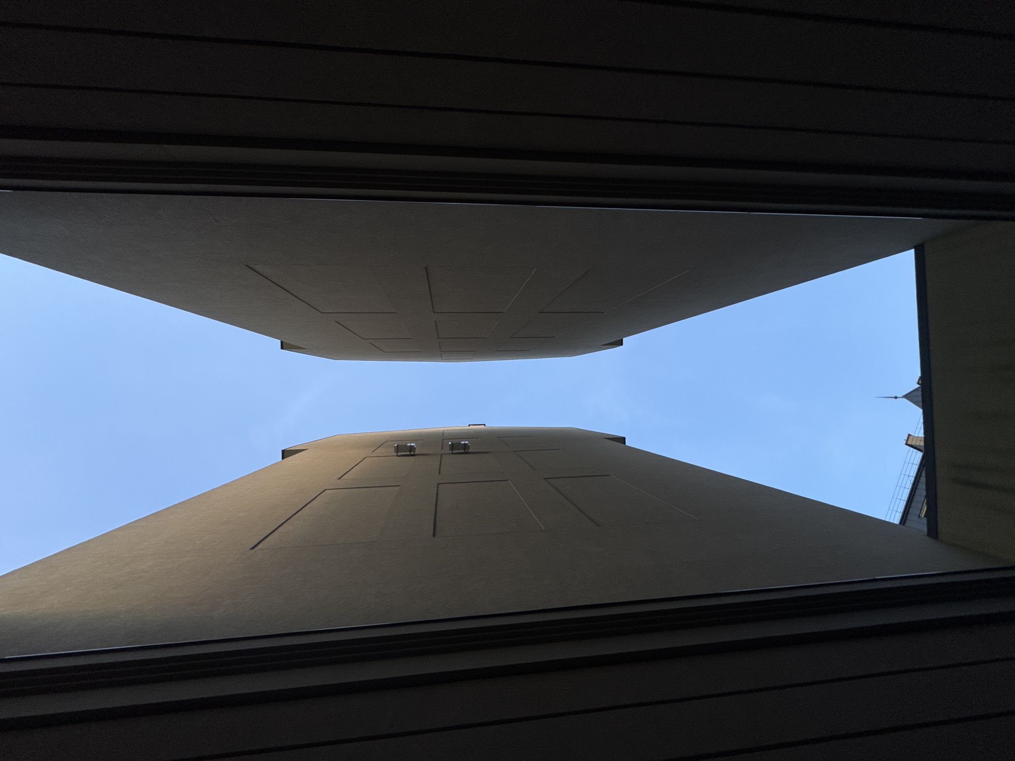 The image presents an upward view of two adjacent buildings in Zelenogradsk, Russia, framed by a dark, likely architectural overhang, possibly part of a window or balcony structure. The buildings are tall, with flat, light-colored facades and simple, rectangular relief patterns. There appear to be small ventilation or service access points on the lower building. The sky is visible between the buildings, and a neighboring roof with metal fencing and a pointed spire is visible on the right. No people are visible. The environment seems to be urban.