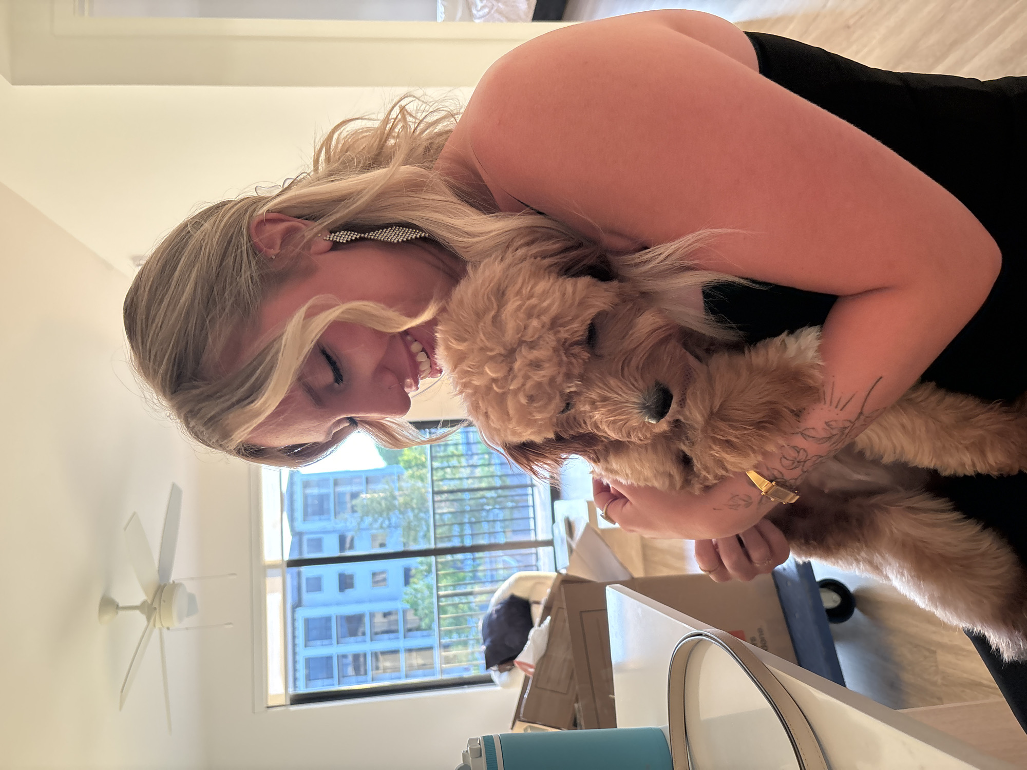 An indoor medium shot captures a fair-skinned woman with blonde hair, wearing a black strapless top and sparkling earrings, embracing a golden-brown, fluffy dog. The woman is looking down at the dog with a gentle smile. Her left arm is wrapped around the dog, holding it close to her chest. A gold wristwatch adorns her left wrist, and she has a ring on her right index finger. The dog appears relaxed, snuggled into the woman's embrace. The background reveals an interior scene with a ceiling fan, a window with a view of buildings and trees, stacked cardboard boxes, and a blue water bottle on a table. The lighting suggests it is daytime. The scene is set in Hallandale Beach, United States of America.