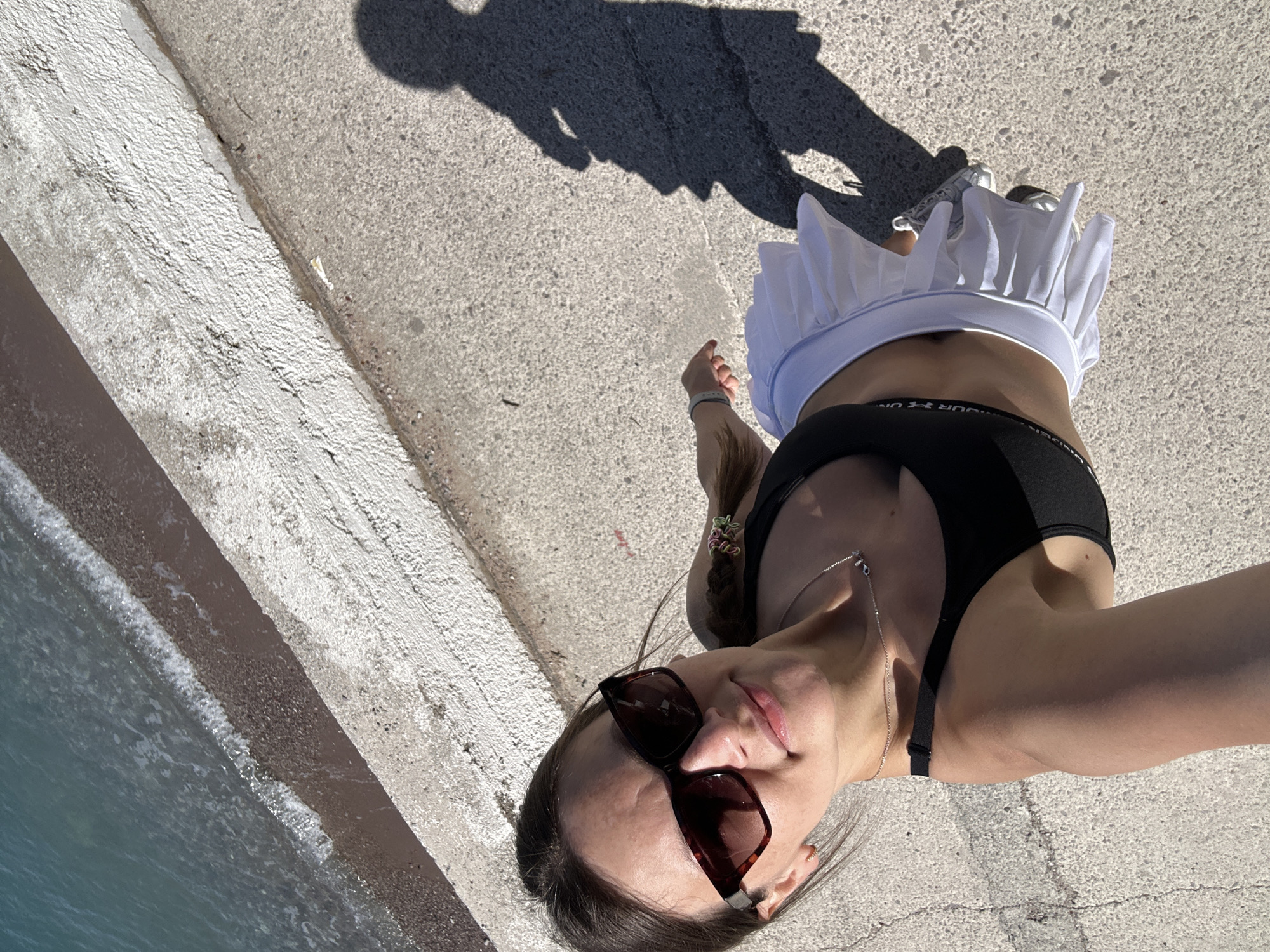 A woman stands on a concrete pier in Boreti, Montenegro. She wears a black sports bra, a white tennis skirt, and sunglasses. A silver necklace hangs around her neck. Her hair is pulled back in a braid. A shadow of a person is cast on the pier behind her. The Adriatic Sea is visible to the left of the pier.