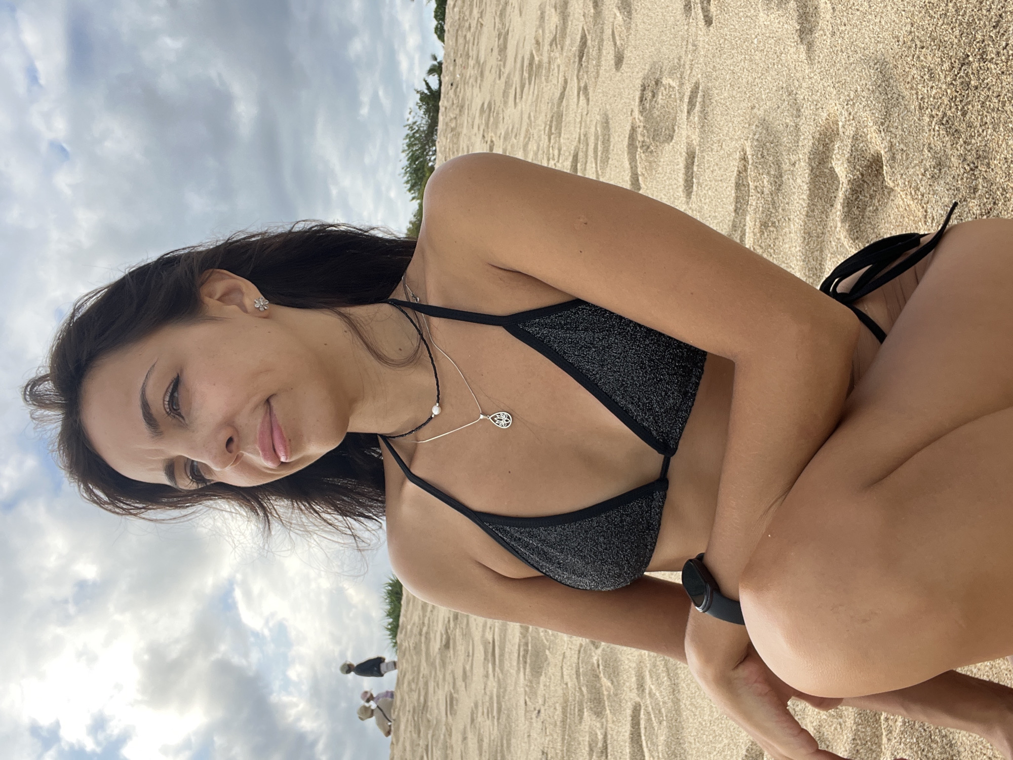 A young woman is crouching on a sandy beach. The sand appears light in color and slightly damp. Behind her is a cloudy sky with a hint of greenery in the background, suggesting vegetation or trees near the beach. There are people standing in the background. The woman is wearing a dark-colored bikini and jewelry including a necklace with a pendant and earrings. A watch is on her wrist. She is looking to the left with a gentle smile. The scene is likely in Kuta Selatan, Indonesia, given the beach setting and the prompt instructions.