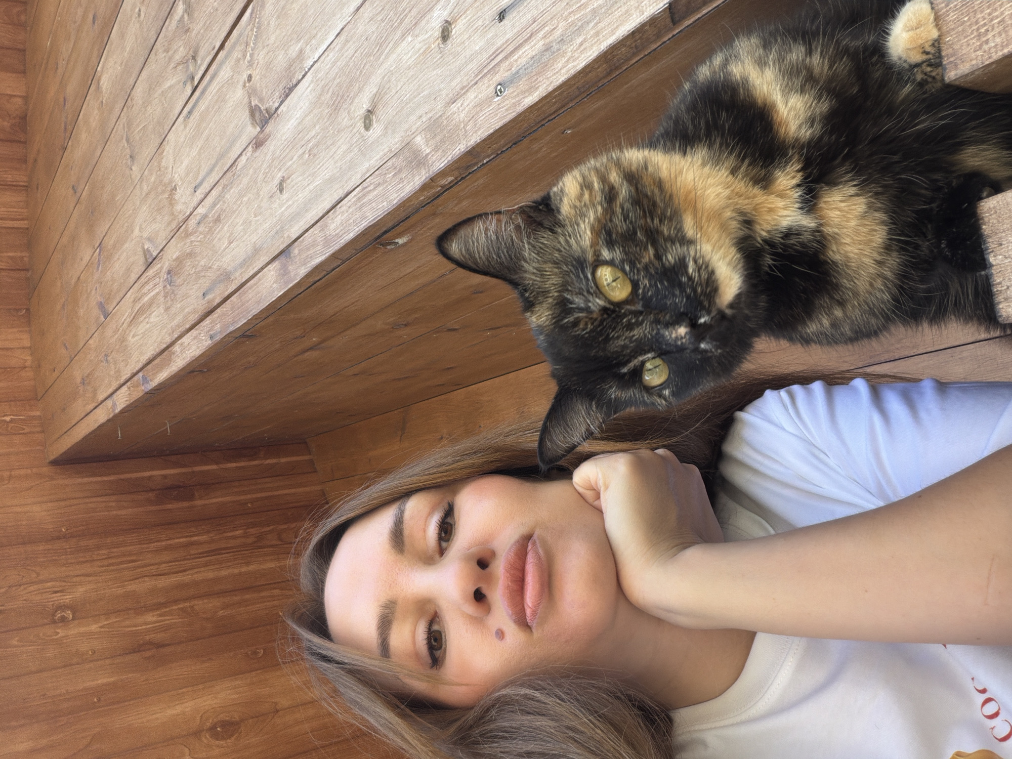 A young Caucasian woman is taking a selfie with a tortoiseshell cat. The woman has long brown hair, is wearing a white t-shirt, and is resting her chin on her hand. The cat is looking directly at the camera with bright yellow eyes. They are both sitting on wooden steps, which appear to be outdoors. The background is also made of wood, likely part of a deck or porch. The setting is likely a residential area in Malinovka, Russia.