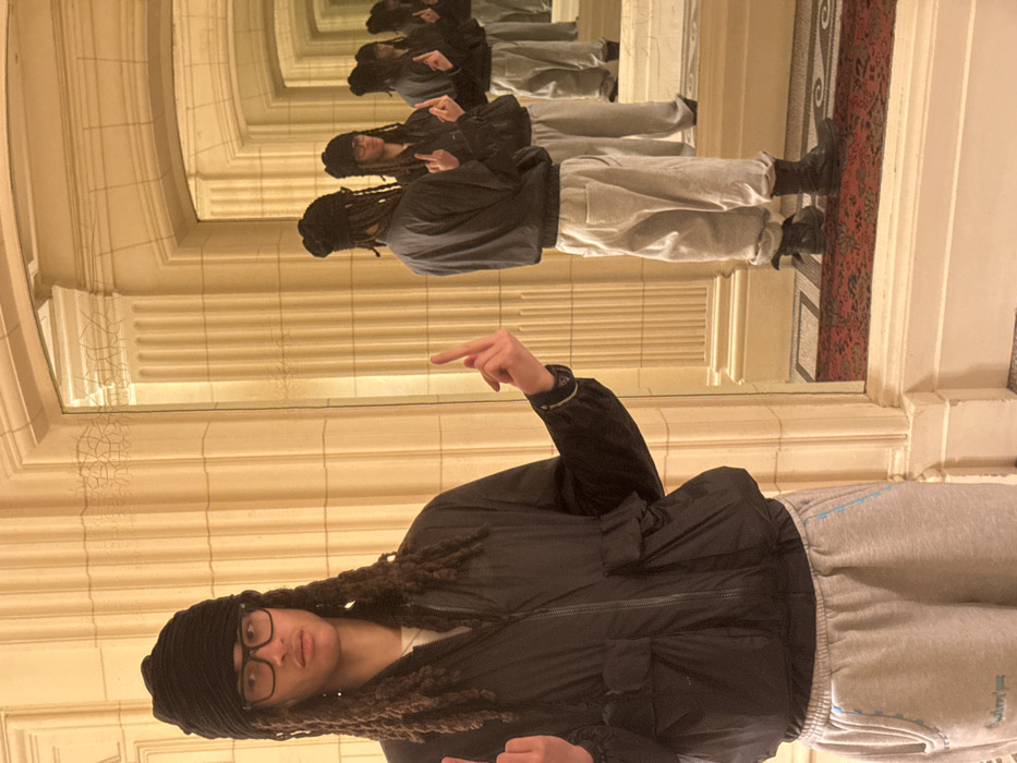 An individual with long dreadlocks, wearing a black knit beanie, black-rimmed glasses, a black zippered jacket, and light grey sweatpants, stands within an ornate interior space in Paris, France. The person faces directly forward, extending the middle finger of both hands. They are positioned before a large mirror, which reflects the individual and the architectural features in a repeating sequence, creating an illusion of infinite depth. The interior features cream-colored or light-toned walls adorned with classical fluted pilasters, decorative cornices, and arched frames around the mirror openings. A red patterned rug with dark motifs covers the floor, visible in the reflections. The lighting is artificial and warm.