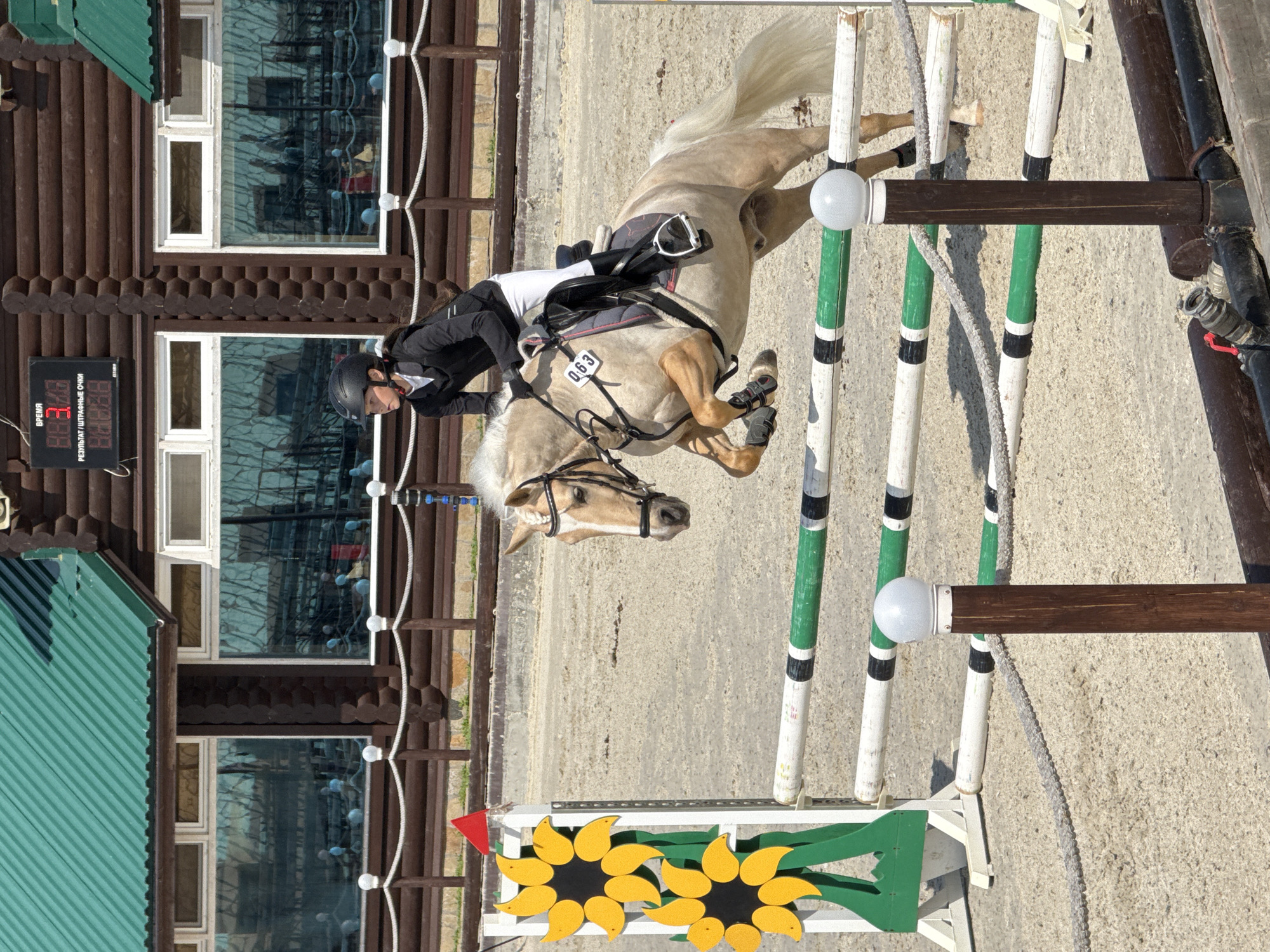 A blond horse and rider are in mid-air, jumping over a white and green equestrian obstacle on a dirt track. The rider, a young female, wears a black riding jacket, white pants, and a black helmet. A numbered bib with "063" is visible on the horse's saddle. In the background, a wooden building with a green roof and large windows stands behind a brown wooden fence. A digital scoreboard displaying "8.8.3.0.8" and "8.8.8.8.8" is mounted on the building. To the left of the obstacle, a decorative element resembling two sunflowers is attached to a white stand. The setting appears to be an equestrian facility, likely in the city of Ferma, Russia.