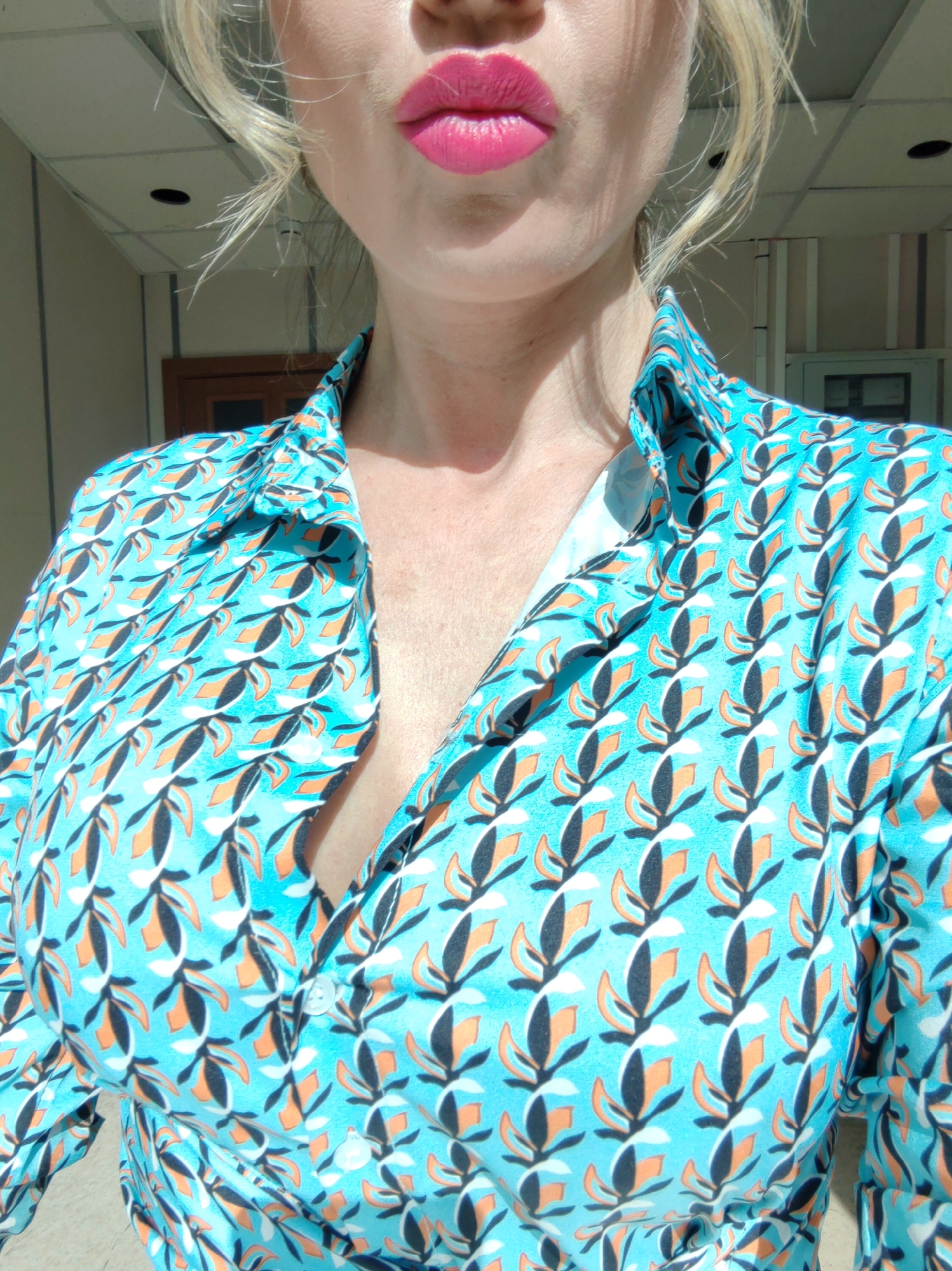 A woman with blonde hair and pink lipstick is wearing a blue, orange, and black patterned button-down shirt. The photo is taken from a slightly low angle, focusing on her face, neck, and the upper part of her torso. Behind her, a doorway and the upper part of a doorframe can be seen, suggesting she is indoors near an entrance. The location is Unknown, Unknown.