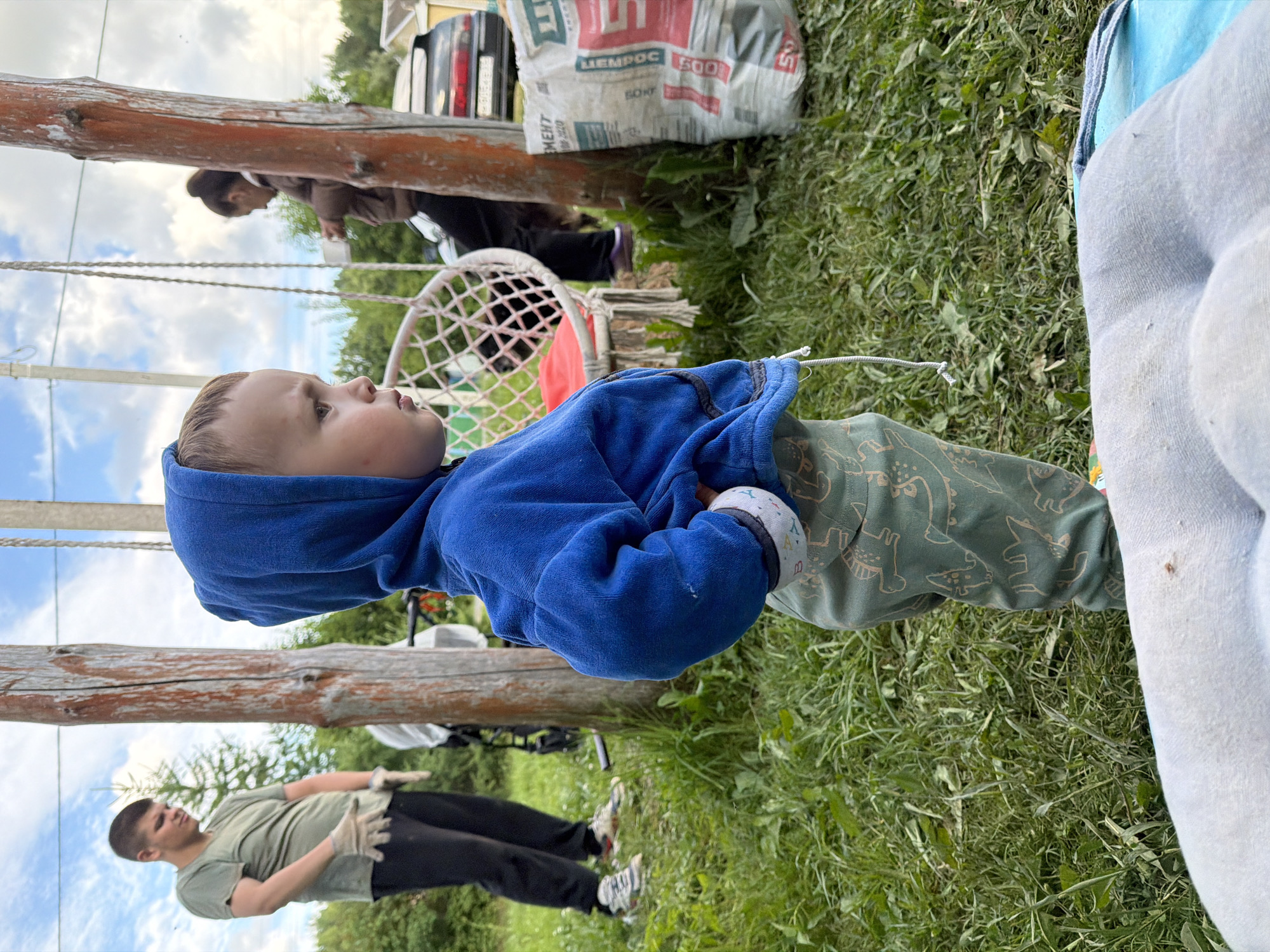 In Красный Маяк, Russia, an outdoor scene unfolds with a young child, likely a toddler, in the foreground wearing a blue hooded sweatshirt and green patterned pants. The child stands with their hands on their hips, facing right. Behind the child, a man in a green t-shirt and black pants, wearing work gloves, is visible, facing left and looking down. Further in the background, a woman stands near a car and a white mesh swing chair. The environment is grassy, with wooden posts supporting some structure, possibly a swing set. A large white bag with red and blue text is also present in the background, near the car. The sky is partly cloudy, suggesting daylight hours.