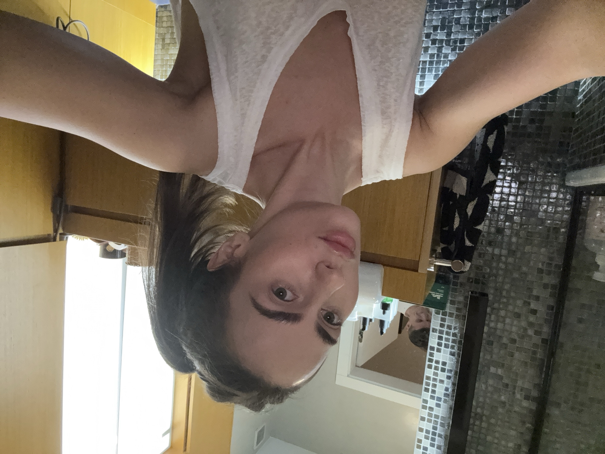 A woman is taking a selfie in a bathroom. She is wearing a white tank top. The bathroom has mosaic tile walls, wooden cabinets, and a mirror reflecting her face. The location cannot be determined to be Warsaw, Poland.