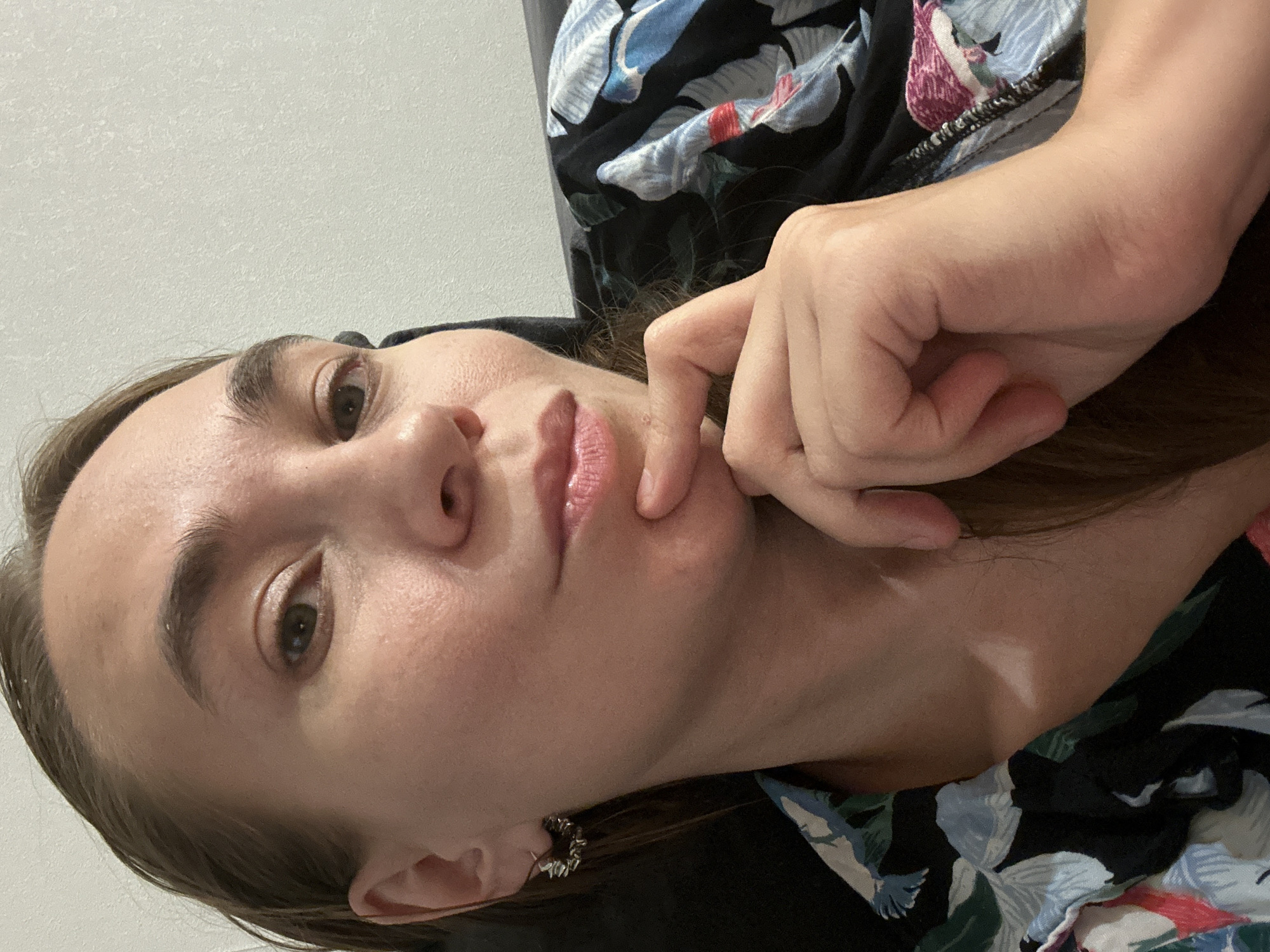A selfie shows a fair-skinned woman with light brown hair and brown eyes, wearing a floral patterned shirt. She rests her chin on her hand. She appears to be indoors, with a plain white wall visible in the background. The location cannot be determined to be Kharnavy, Russia, based on the image.