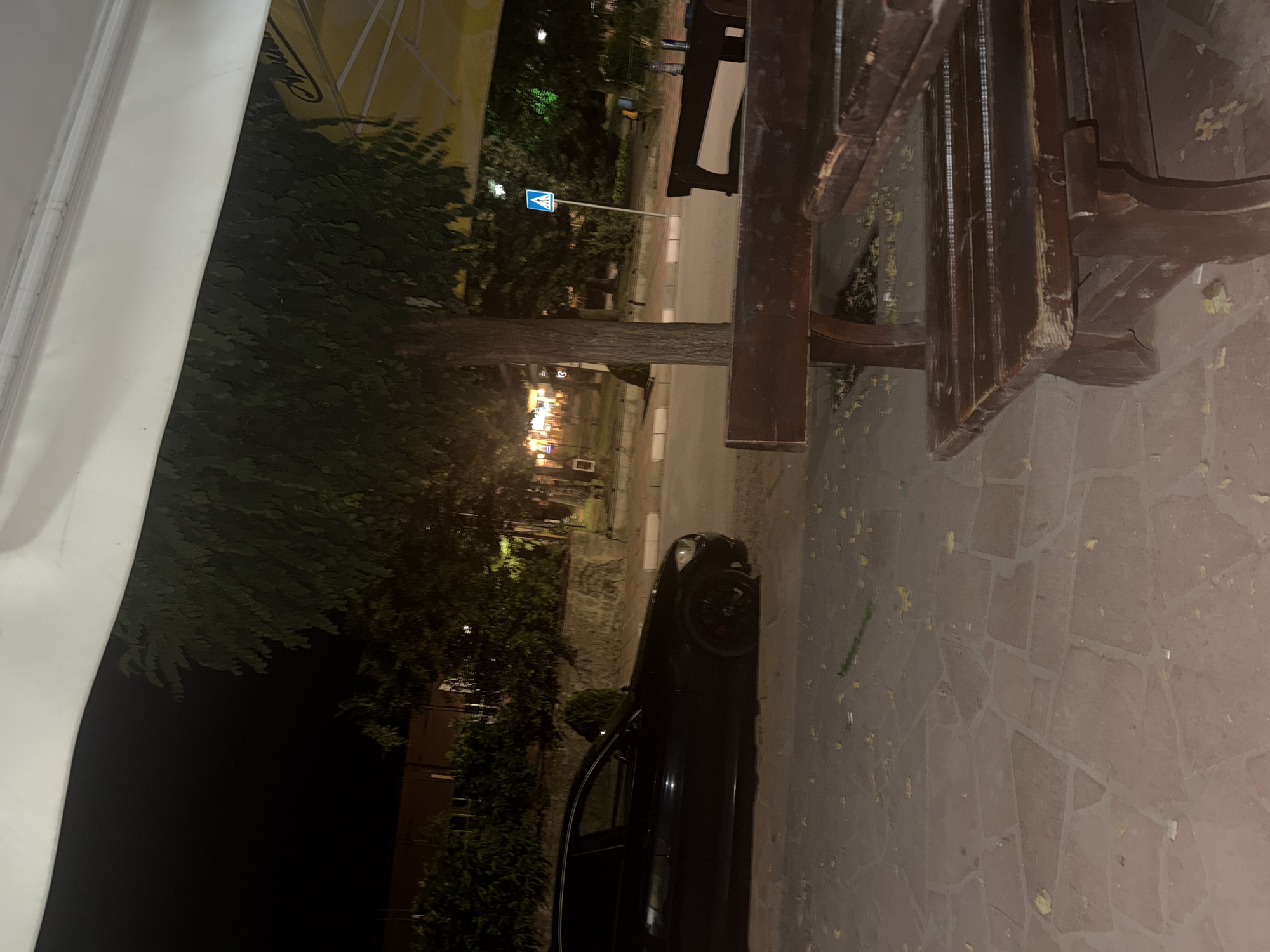 The image depicts an outdoor scene at night, possibly a sidewalk cafe or restaurant seating area. In the foreground, a dark wooden picnic table and bench are visible, showing signs of wear. The ground is paved with irregular-shaped stones, and scattered fallen leaves are present. To the left, a black sedan is parked. Beyond the seating area and car, a street is visible with a pedestrian crossing sign. Trees and some building lights can be seen in the background, suggesting an urban or suburban setting. The overall lighting is dim, characteristic of nighttime.