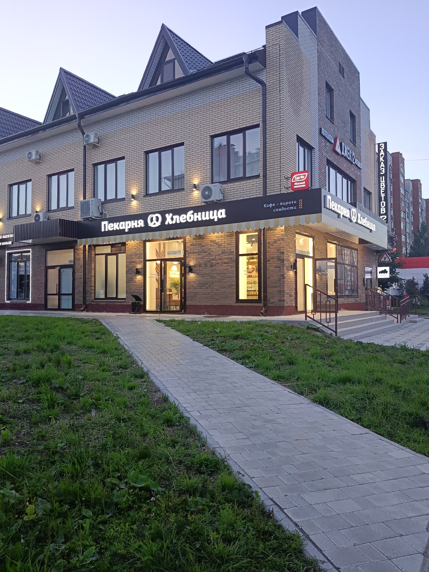 This media file shows a commercial building with businesses on the ground floor. The building is constructed of light-colored brick and has a sloping dark roof. Several air conditioning units are mounted on the facade. The businesses have illuminated signs. The largest sign reads "Пекарня Хлебница" (Bakery Khlebnitsa), and another sign indicates "Кофе пироги сладости" (Coffee, pies, sweets). Another sign reads "Tiena Мясной магазин" (Tiena Meat store). To the right, a tall vertical sign displays "3AKA3 UBETOB" (Order flowers).
In front of the building, a paved walkway leads from the street towards the entrance. There is a grassy area with weeds and some plants to the left of the walkway. The building's windows are lit from within, suggesting it is evening or early morning. The sky is a pale blue, indicating either dawn or dusk. The overall scene is quiet and appears to be in a residential or mixed-use area, given the adjacent apartment building visible on the right side of the image. There are no people visible in the scene.