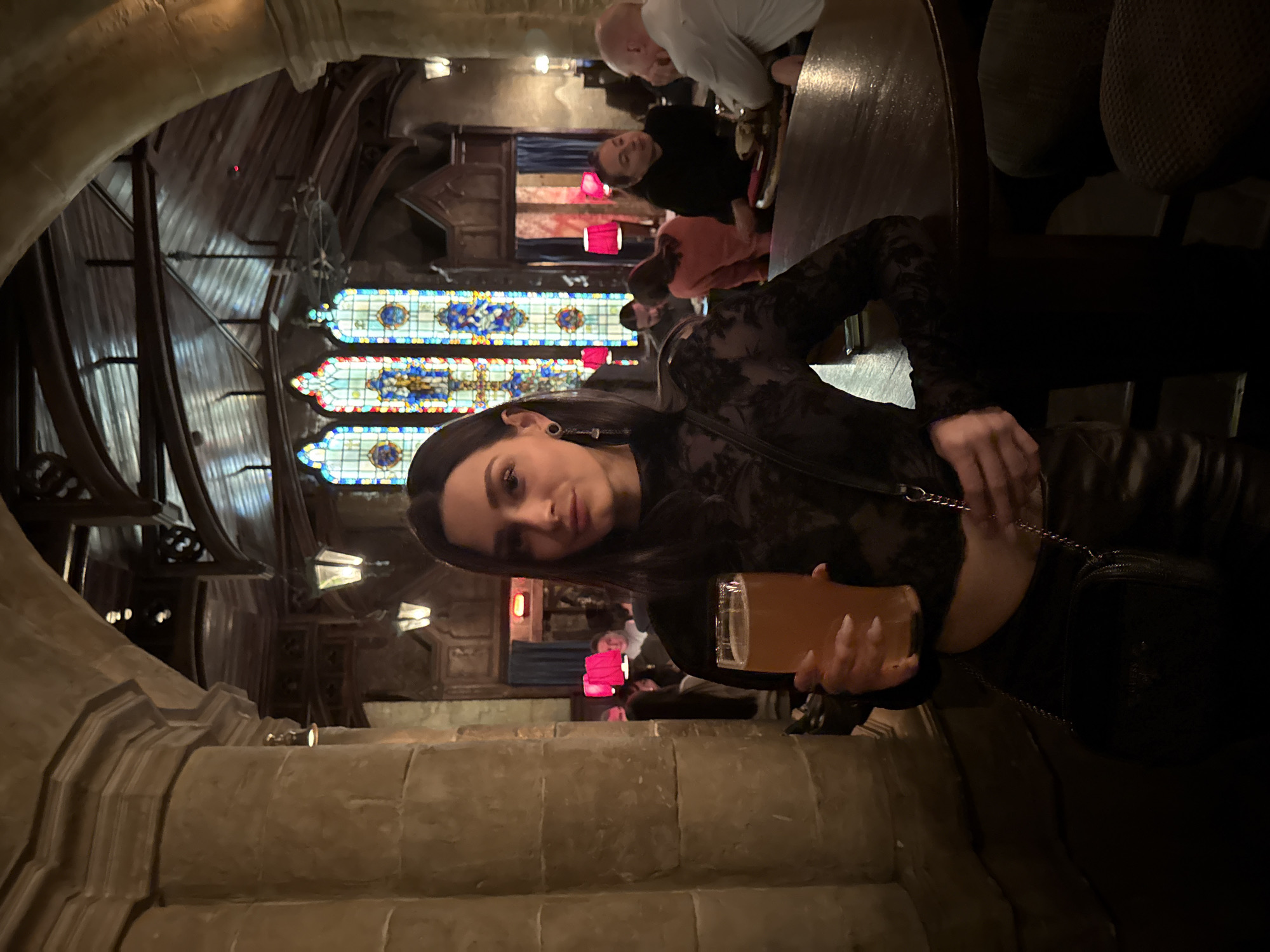 The image shows a young woman holding a glass of beer in what appears to be a restaurant or pub. The background includes stained glass windows, arched ceilings, and stone structures, suggesting a historical or themed establishment. Other patrons can be seen in the background. The location appears to be the Okhotny Ryad shopping mall in the city of Moscow, Russia.