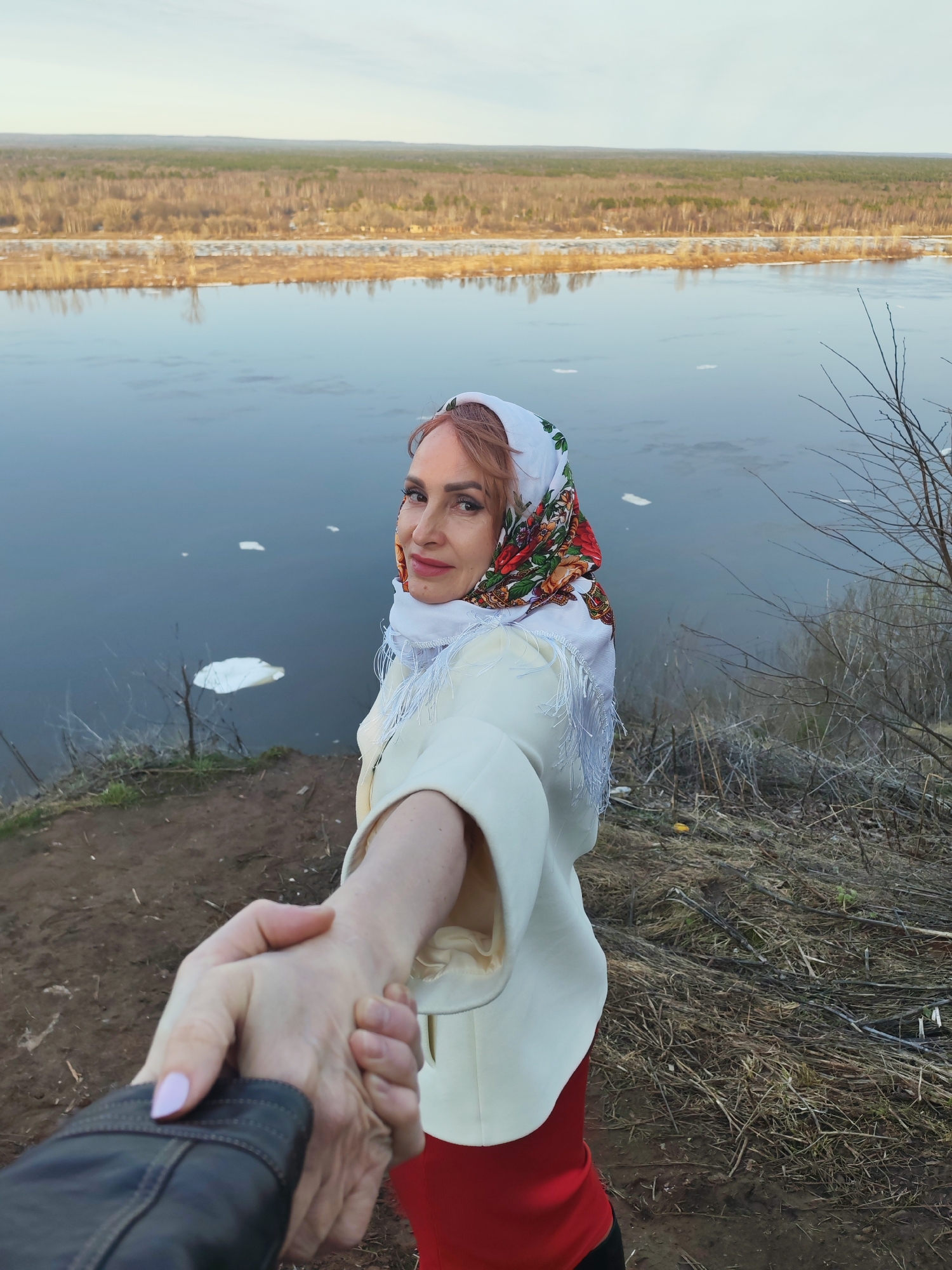 A person with reddish hair is holding another person's hand on a cliff overlooking the Kama River in Sarapul, Russia. The person is wearing a white headscarf with a floral pattern, a white jacket with fringe, and a red skirt. The person holding the hand is wearing a black leather jacket and has pale pink nail polish. The river is wide and has ice floes floating in it. The opposite bank of the river is visible, with a forest and buildings in the distance. The cliff is covered in dry grass and some bare trees. The sky is overcast.