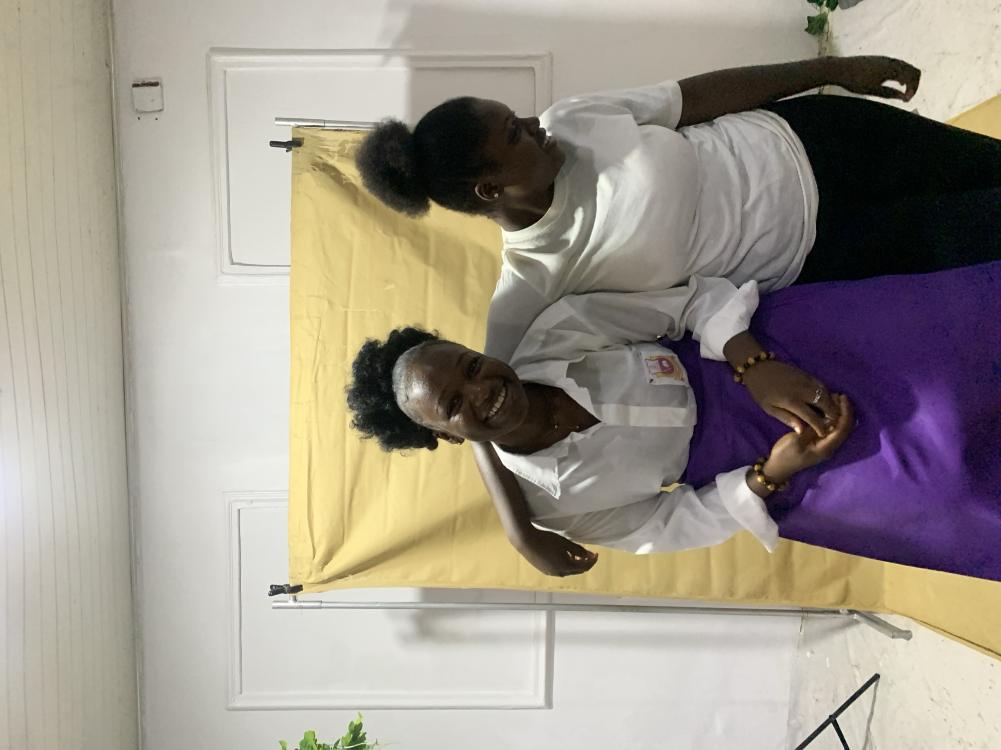 Two Black women are posing for a photo in front of a yellow backdrop that is hung in front of a white wall, in a location that is likely in Jos, Nigeria. One woman wears a white t-shirt and black pants and is lying on her side with her head near the top of the frame. The other woman is wearing a white button-down shirt and a purple skirt, and she is seated. The standing woman has her arm around the seated woman's shoulder, and they are both smiling. The environment includes a white wall and baseboard, a white door frame, and a green plant is located to the side of the image.