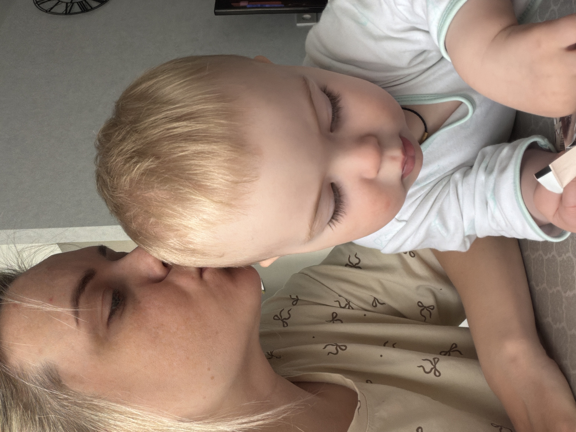 This media file captures a tender and intimate moment between a woman and a young child in an indoor setting, likely Votkinsk, Russia.
The primary subjects are a woman, presumably the mother, and a baby or toddler. The woman has blonde hair and fair skin with visible freckles. Her eyes are closed or looking down, and her face expresses a sense of peace and affection. She is wearing a light-colored top patterned with small, dark brown bows or ribbons.
The child, also with blonde hair, is resting their head gently on the woman's forehead or nose, with their eyes closed, suggesting they are sleeping or in a deeply relaxed state. The child is dressed in a white t-shirt or onesie with light blue trim. A dark beaded necklace is visible around the child's neck, and they appear to be holding a small, light-colored object in their right hand.
The activity depicted is a quiet moment of rest or sleep, highlighting the close bond and comfort shared between the mother and child. The overall emotion conveyed is one of profound love, peace, and maternal tenderness.
In the background, a large, ornate wall clock with Roman numerals is partially visible, suggesting a domestic environment. Part of a patterned cushion or blanket can also be seen. The lighting appears to be natural daylight. There is no visible text in the image.