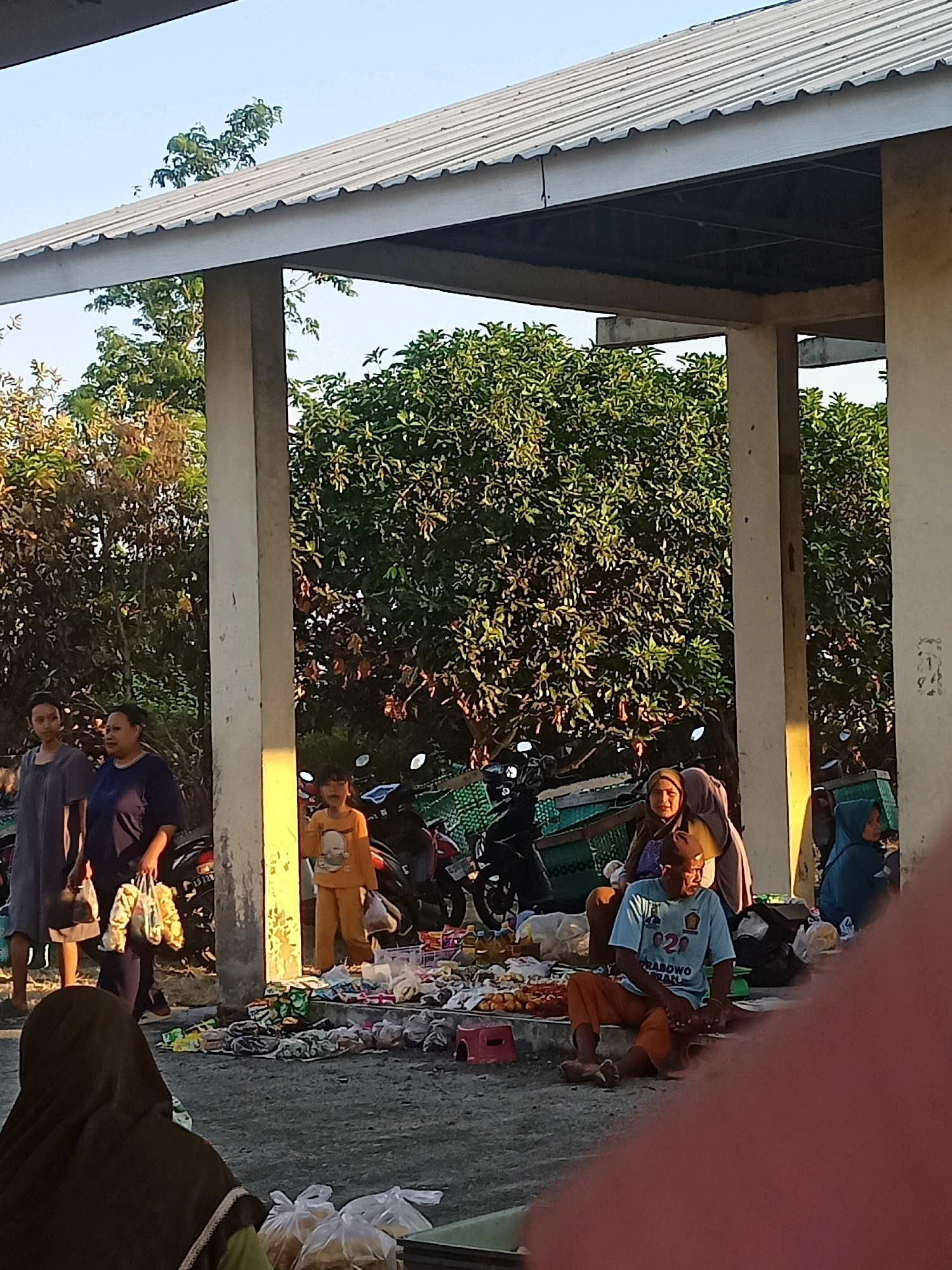 This image captures a scene at a market in Nganti, Indonesia, under a covered structure with a corrugated metal roof supported by concrete pillars. Sunlight casts shadows and highlights the area. Several people are present, some standing and others sitting. In the foreground, a person wearing a dark hijab is seated, with plastic bags of goods in front of them. To the right, a man in a light blue t-shirt and orange pants is seated, with more goods laid out around him. A woman wearing a headscarf is seated behind him. Further to the left, two women are standing, one in a dark blue dress and the other in a navy blue top, holding shopping bags. A young child in an orange outfit stands near them, also holding a plastic bag. Several motorcycles are parked behind the people and merchandise, suggesting transportation. The ground is paved, and various items are displayed for sale, including food items like fruit and packaged goods, as well as other merchandise. Trees and foliage are visible in the background, indicating an outdoor market setting within a populated area.