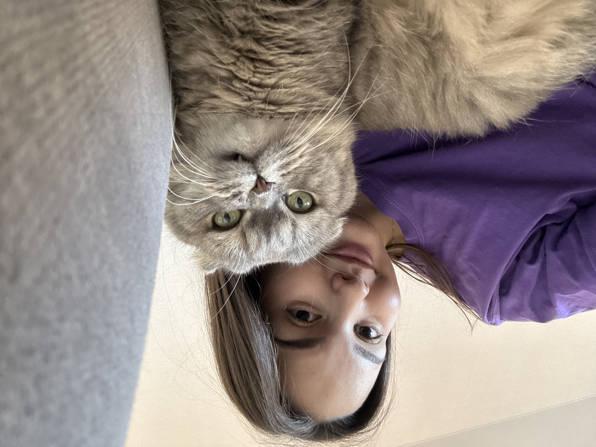 A woman and a gray tabby cat are positioned together in a close-up, upside-down selfie. The woman is wearing a purple shirt. The background is blurred but appears to be a light-colored surface, likely a floor or table. The image does not provide information regarding the city of Chaikovsky, Russia.
