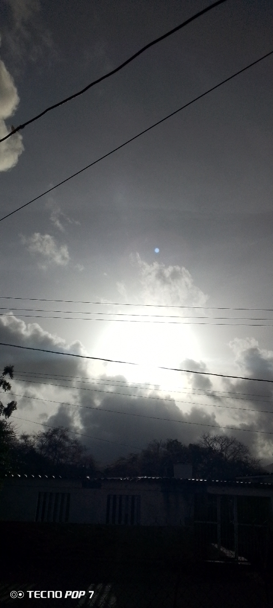 The image shows a daytime scene in Altagracia, Venezuela. The sky is overcast with thick clouds, and the sun is partially obscured, creating a bright glare. Several electrical or telephone wires stretch across the sky. Below the sky is a line of trees and foliage silhouetted against the bright sky. At the bottom of the frame is the dark outline of a building, likely a house, with visible windows. The bottom left corner of the image has the watermark "TECNO POP 7," indicating the device used to take the picture.