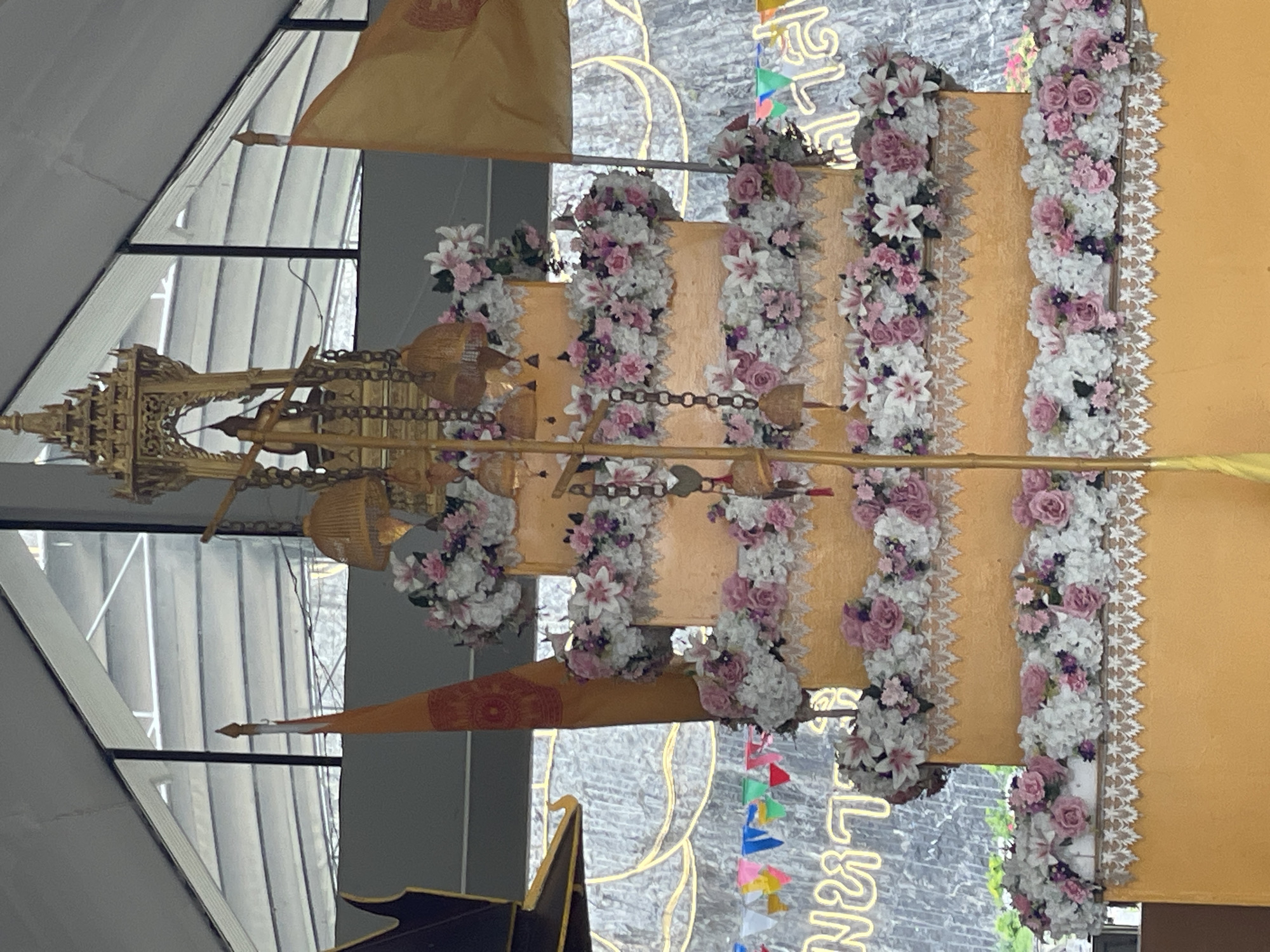The image shows a large, tiered, orange-colored structure adorned with floral arrangements, resembling a cake or monument. The floral decorations consist of white and pink flowers. Atop the structure, a golden, ornate canopy holds a small statue of a figure, possibly a Buddha or a deity. Several golden bells hang from chains suspended from the canopy. Two flags are visible on either side of the structure; one is orange with a symbol and the other is yellow. In the background, a rocky or concrete wall is visible, with decorative yellow lettering and a string of colorful triangular flags. The overall scene appears to be outdoors, possibly within a temple or a public space in Tambon Bang Sare, Thailand, given the context provided. The lighting suggests it is daytime. There are no visible people in the image.