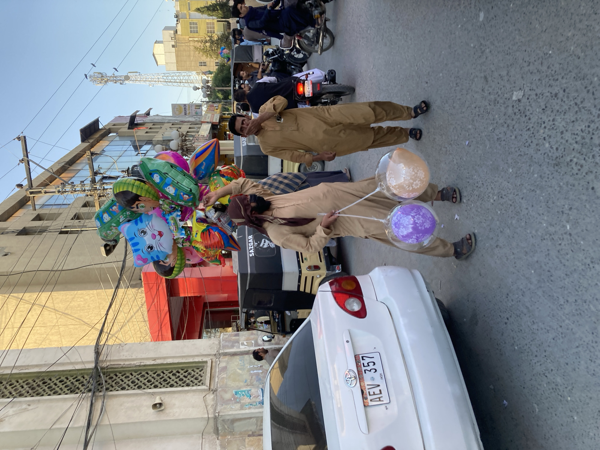 Two men stand on a street in Quetta City Tehsil, Pakistan. One man wears a brown shalwar kameez and holds a bundle of colorful character balloons, while the other wears a similar outfit and has his hand near his mouth. A white car with license plate "AEV-357" is partially visible. In the background, there are motorcycles and an auto rickshaw. The surrounding buildings suggest an urban setting.