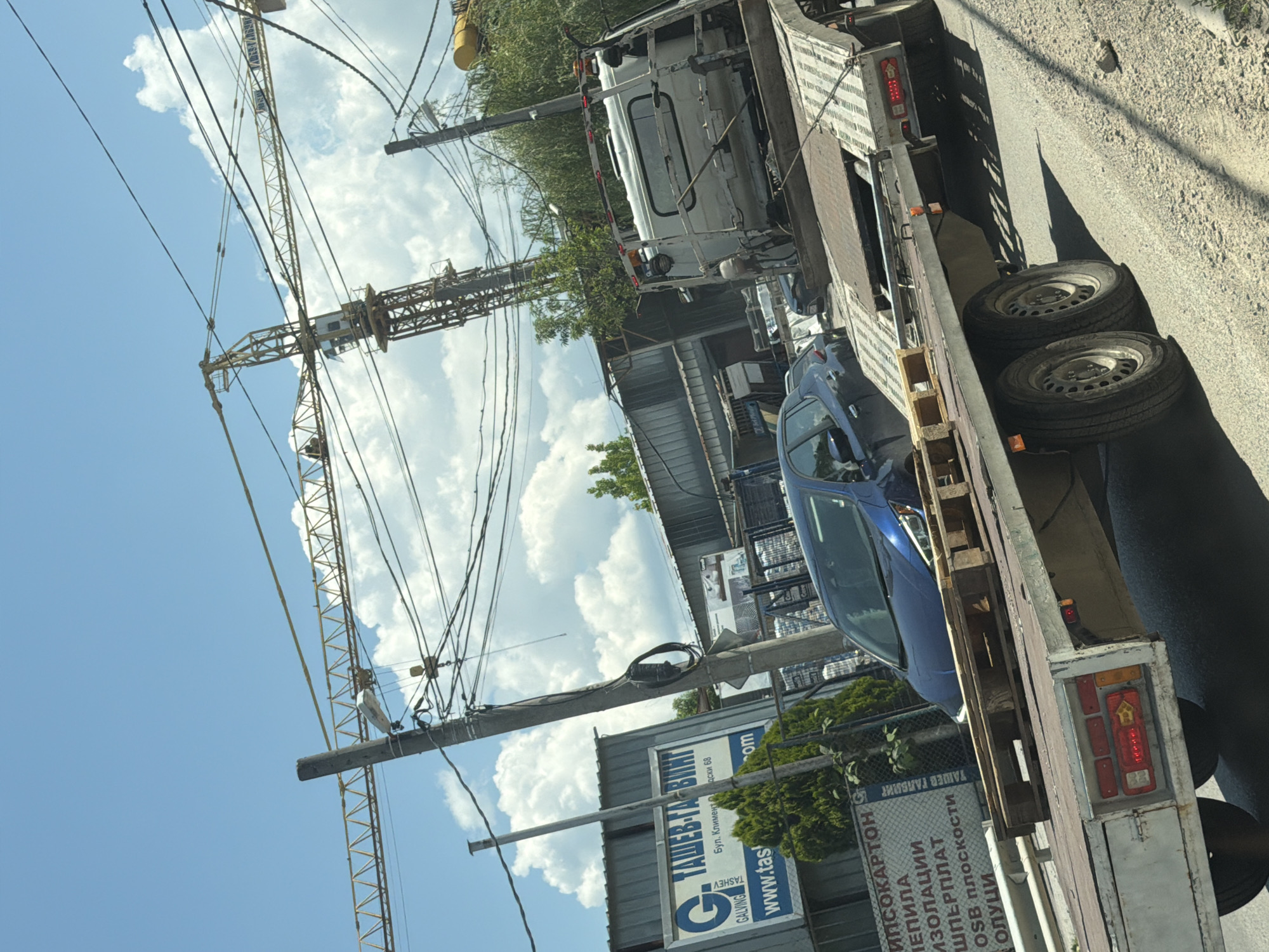In an outdoor, urban setting, likely in Sofia, Bulgaria, a flatbed tow truck is parked on the side of a road. The tow truck is carrying a blue sedan and several wooden pallets on its bed. Behind the tow truck, a row of industrial buildings with corrugated metal siding is visible. Overhead, a complex network of electrical wires crisscrosses the sky, supported by utility poles. A construction crane, a prominent structure, is positioned in the background. The sky is blue with scattered white clouds, and the sun is casting shadows. The ground appears to be asphalt and gravel. A sign in Bulgarian, advertising construction materials like plasterboard, insulation, and OSB boards, is attached to one of the buildings.
