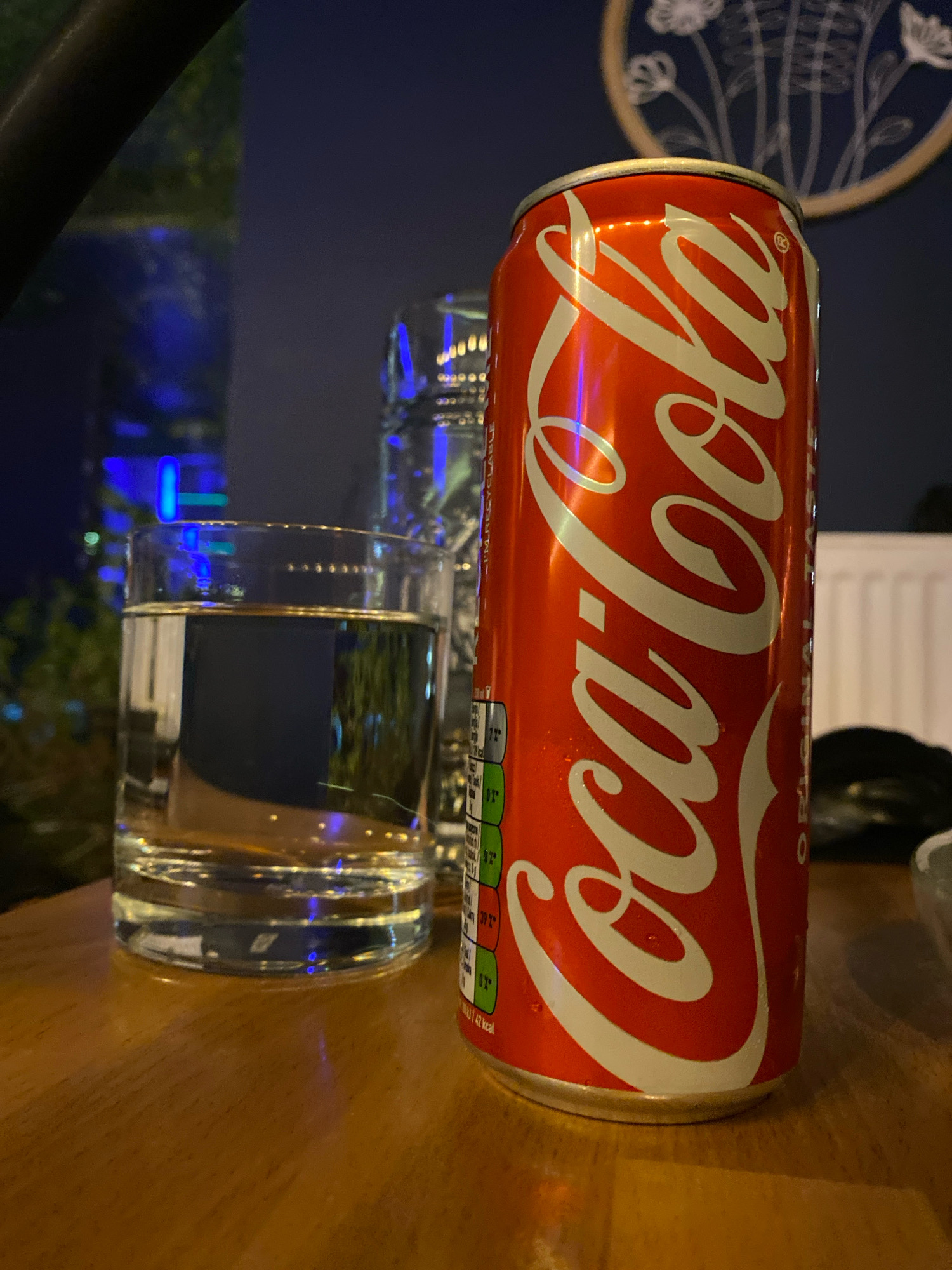 A close-up, eye-level shot shows a red Coca-Cola can with the iconic white logo prominently displayed. The can is slightly angled towards the viewer and appears to be sitting on a wooden surface. To the left of the can is a clear glass filled with a light-colored liquid, likely water. Behind the glass, another clear container, possibly another glass or bottle, is visible but out of focus. The background is dark and blurry, with hints of blue and green light, suggesting an indoor setting with ambient lighting, possibly a restaurant or bar. A decorative plate with white floral motifs on a dark blue background is visible on the far wall. The scene does not contain any people or identifiable landmarks that would allow for specific location identification within Gliwice, Poland.