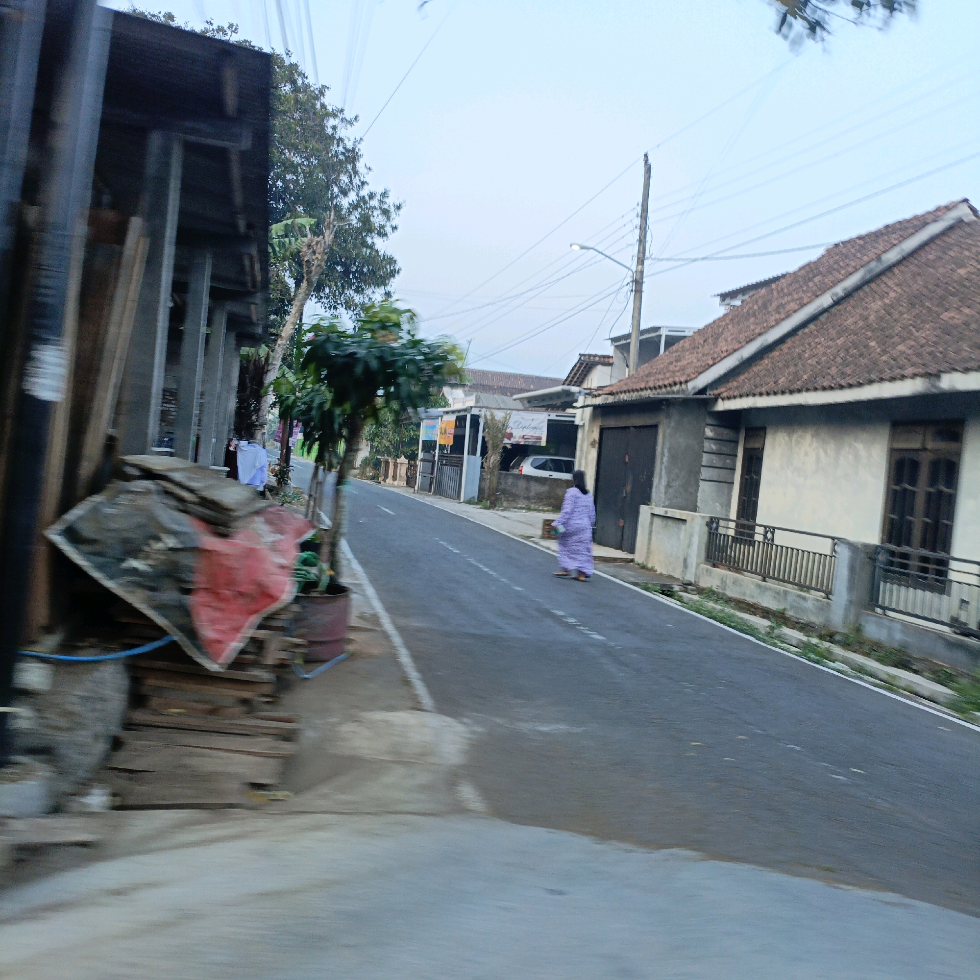 The image captures a street scene in Salatiga, Indonesia, with a focus on a residential area. A woman, dressed in a long purple garment, is walking away from the camera on the asphalt road. To the right of the road, a house with a terracotta tiled roof and light-colored walls is visible, featuring wooden-framed windows and a dark wooden door. A low concrete wall with a metal railing runs along the front of the house. Across the street, on the left side of the image, there's a structure with wooden beams and corrugated metal siding, appearing to be a covered storage area or an unfinished building. Piles of wood and what looks like a red tarp are stacked in front of this structure. A small tree in a pot stands near the curb, and another larger tree with green foliage is further down the street. Utility poles with numerous wires are scattered throughout the background, indicating the presence of electrical and communication infrastructure. The sky is overcast and pale. The overall impression is a quiet, possibly residential street in an Indonesian town or city.