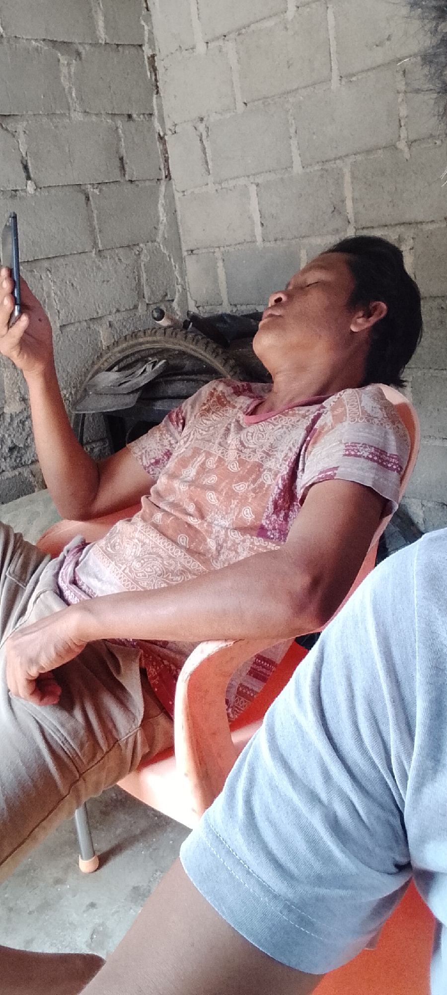 In Padang Dalom, Indonesia, a person is seated in a plastic chair, holding a mobile phone in their right hand and looking at it. The person is wearing a patterned shirt and beige pants. The background consists of a brick wall and a bicycle wheel. Another person's arm is visible in the foreground, wearing a light blue t-shirt. The scene appears to be outdoors, possibly in a yard or an open-air space.