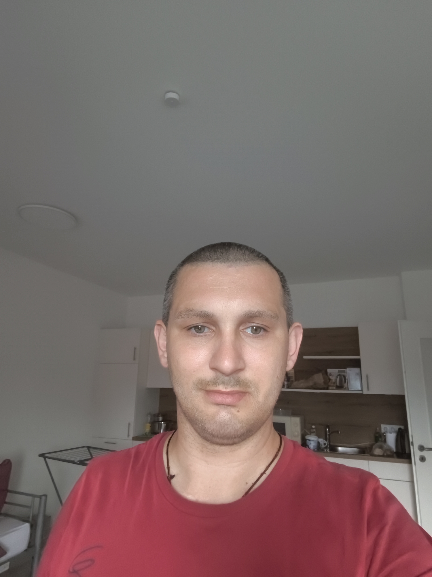 A man with short-cropped hair and a light beard is taking a selfie in what appears to be a kitchen or living area. He is wearing a red t-shirt and a dark necklace. Behind him, a kitchen counter with cabinets, a sink, and some appliances is visible. To the left, a drying rack and part of a chair are in the foreground. The ceiling has a light fixture and a smoke detector. This scene is located in Wolgast, Germany.