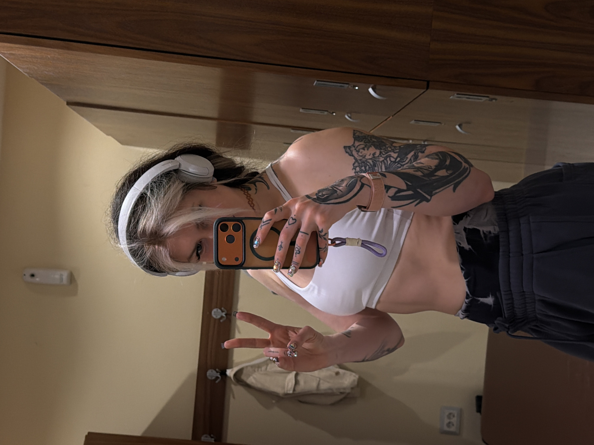 A young woman stands in front of a mirror, taking a selfie. She is wearing headphones and a white sports bra. Her left arm is extensively tattooed. She is holding a cell phone in her right hand. She is making a peace sign with her left hand. The reflection in the mirror shows the background, including a wooden dresser, a wall with a coat rack, and an electrical outlet. The location appears to be an interior space, potentially a hotel room in Budva, Montenegro.