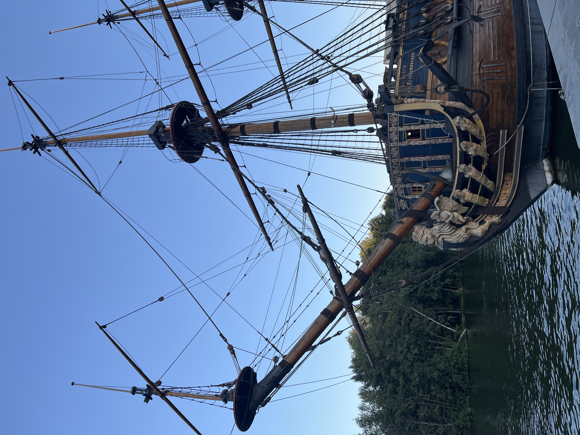 The image shows the bow of a large wooden sailing ship docked on a body of water. The ship is elaborately decorated with carved figures, including lions wearing crowns, and has multiple masts with rigging. The hull is painted in dark blue and wood tones. In the background, there is a dense line of green trees on the shore, and a clear blue sky above. The ship is identified by lettering on its side as "Вд-02-0352 Предestinация", and it is located in Novaya Usman, Russia. There are no people visible in the image.