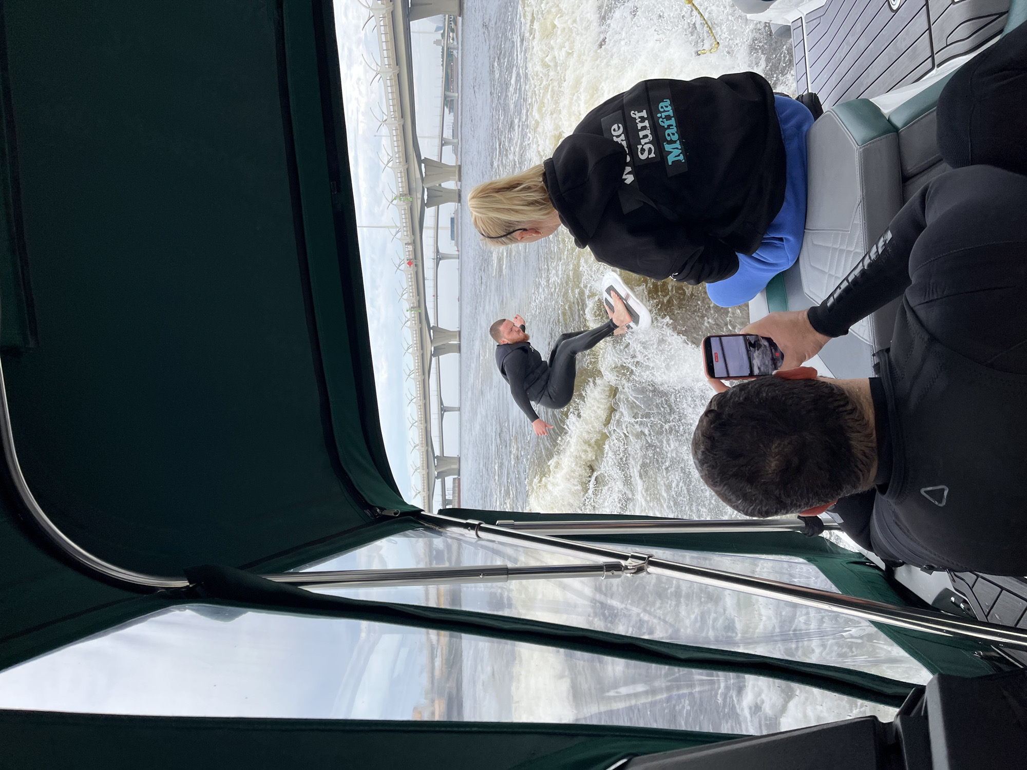A man in a wetsuit is wakeboarding on a river, being pulled by a boat. Another person in a wetsuit is sitting on the back of the boat, holding a cell phone to take a picture of the wakeboarder. A third person, also wearing a wetsuit, is sitting on the back of the boat and holding a cell phone to record the wakeboarder. The boat has a dark green canopy. In the background, a large bridge spans the river. The scene takes place on the Neva River in Saint Petersburg, Russia. The bridge in the background is likely the Western High-Speed Diameter bridge.