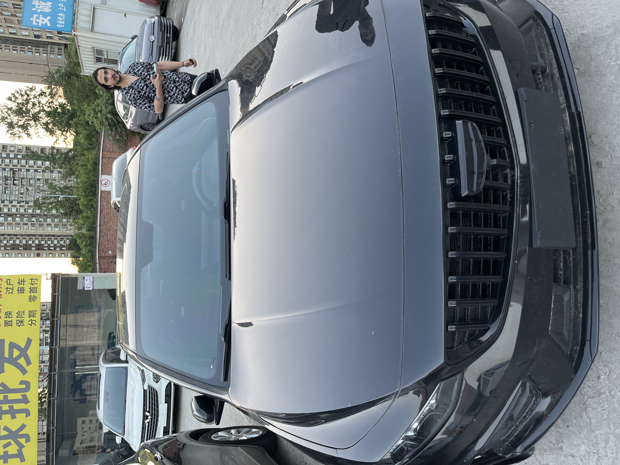 Here is a description of the image:
A slightly rotated overhead shot captures the front end of a dark-colored car. The focus is sharp on the car's grill, headlights, and hood, which reflects ambient light and outlines. The hood has a reflection of a person.
To the left of the car, a man stands near another car, seemingly gesturing towards the camera. In the background are buildings, suggesting an urban environment.
To the lower left is a yellow sign with Chinese characters, attached to a structure. Other cars are visible in the background.
Based on the signs, the location is Banan, China.