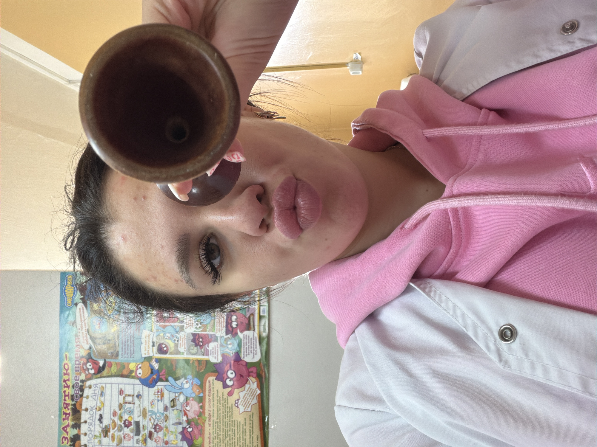 A young woman with dark hair and light skin is taking a selfie indoors. She is wearing a pink hooded sweatshirt under a white lab coat. She is holding a brown, conical object to her eye, obscuring part of her face. Her lips are pursed. In the background, a colorful educational poster with cartoon characters is visible on a wall. The wall behind the woman is yellow. There are no visible geographical markers to indicate a specific location within the city of Chumna, Russia.