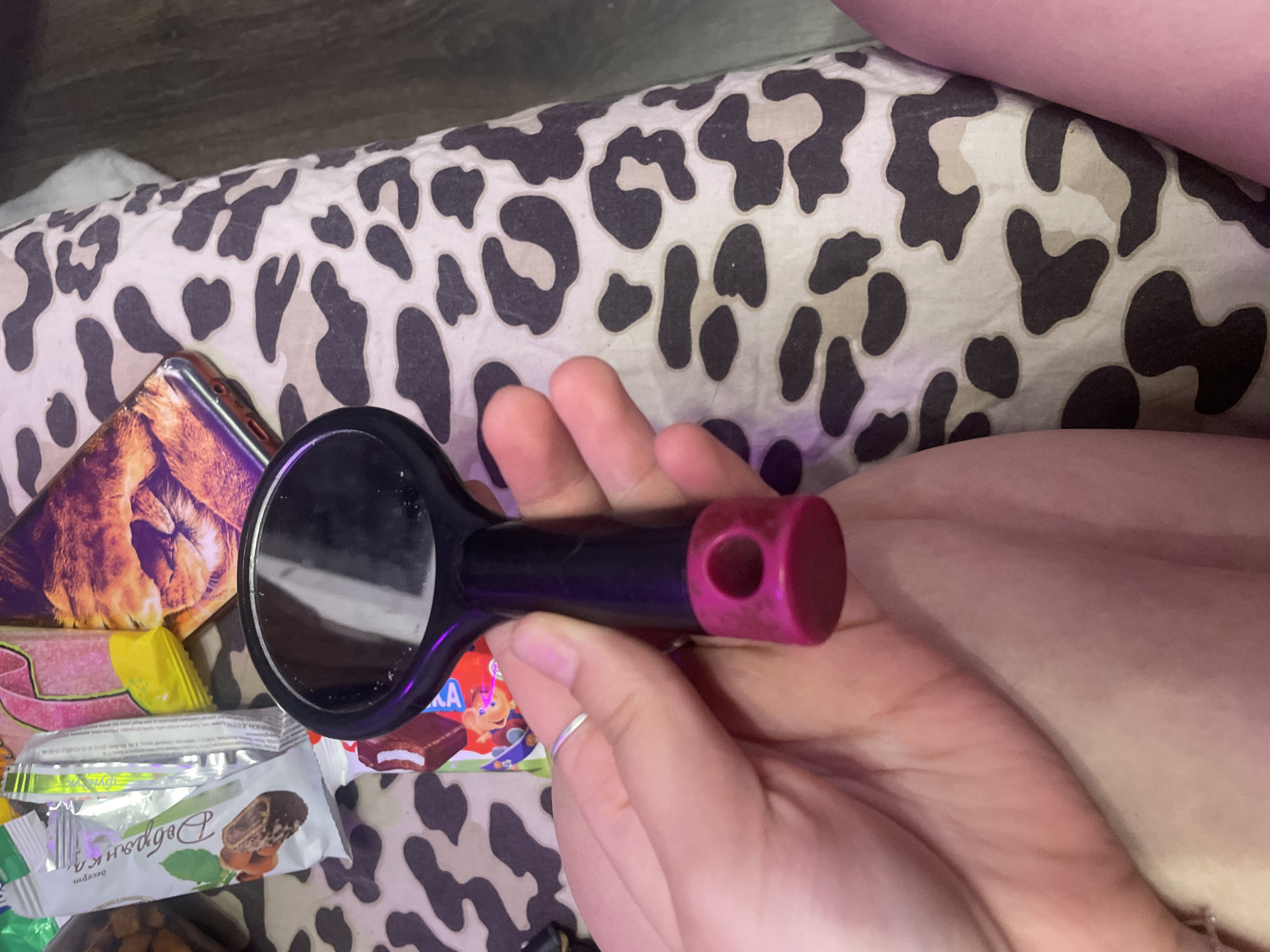 The image shows a close-up of a person's hand holding a small black and pink hairbrush. The background is a patterned fabric that resembles leopard print. Several wrappers of confectionery items are visible in the upper left corner of the image, including a white wrapper with green and brown accents, and a pink and yellow wrapper. There is also a glimpse of a mobile phone with an image of a cat on its case. The lighting suggests an indoor setting, possibly a bedroom. The image does not provide any information about the location in the city of Kornevo, Russia.