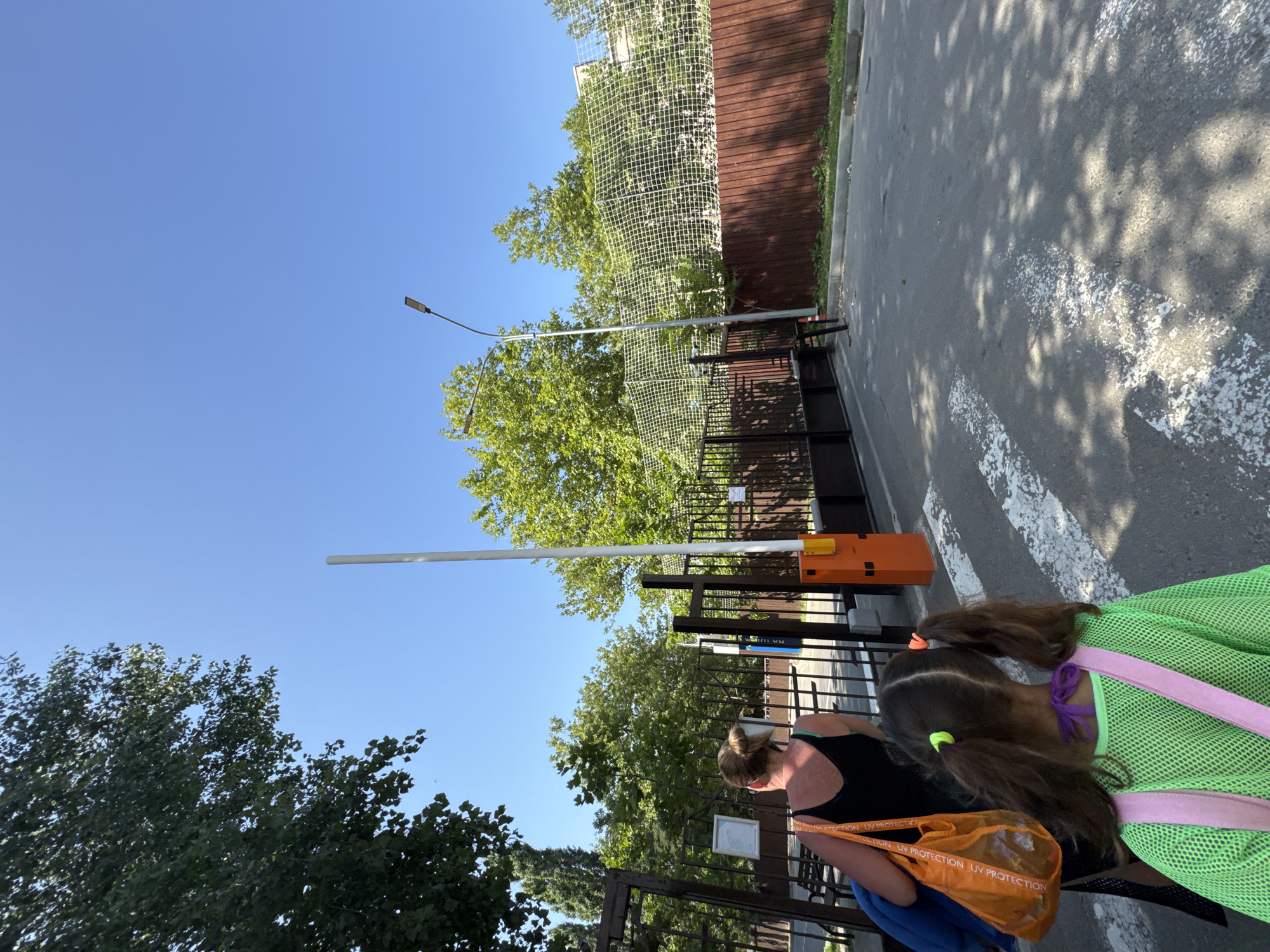 In Vityazevo, Russia, on a sunny day, two individuals, a woman and a young girl, walk towards a closed gate. The woman, seen from the back, wears a black tank top and carries an orange bag over her shoulder, which contains a blue item. She has her hair tied up in a bun and her shoulders appear sunburned. The young girl, also facing away, has her hair in two pigtails with neon green and orange ties. She wears a neon green mesh shirt and a purple ribbon around her neck, with pink straps visible. They are walking on an asphalt road marked with white pedestrian stripes. To their right, a tall white barrier gate with an orange base stands closed, suggesting restricted access. Beyond the gate, a dark metal fence stretches across the scene, with trees and foliage visible behind it. Further back, a brown wooden fence is topped with a white net, possibly a sports court or enclosure. A street lamp with a curved arm extends from the right side of the frame, and a mature tree with lush green leaves shades a portion of the road. The sky is clear and bright blue.