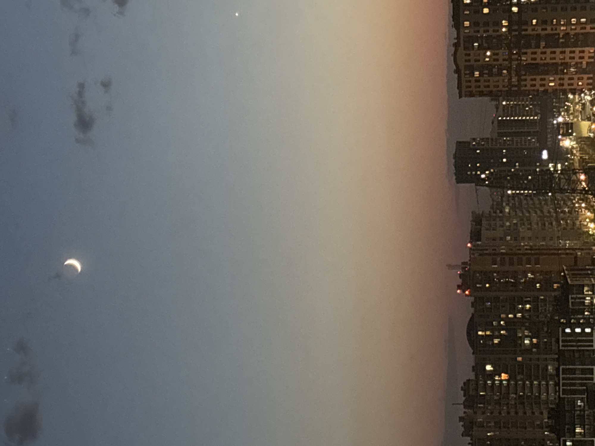 The image shows a cityscape at night with a crescent moon in the sky. The moon is on the left side of the frame, and the cityscape is on the right. The sky is a gradient of blue and orange. The buildings are tall and dense, with many lit windows. A small, bright star or planet is visible near the top of the frame. The image appears to be rotated 90 degrees clockwise.