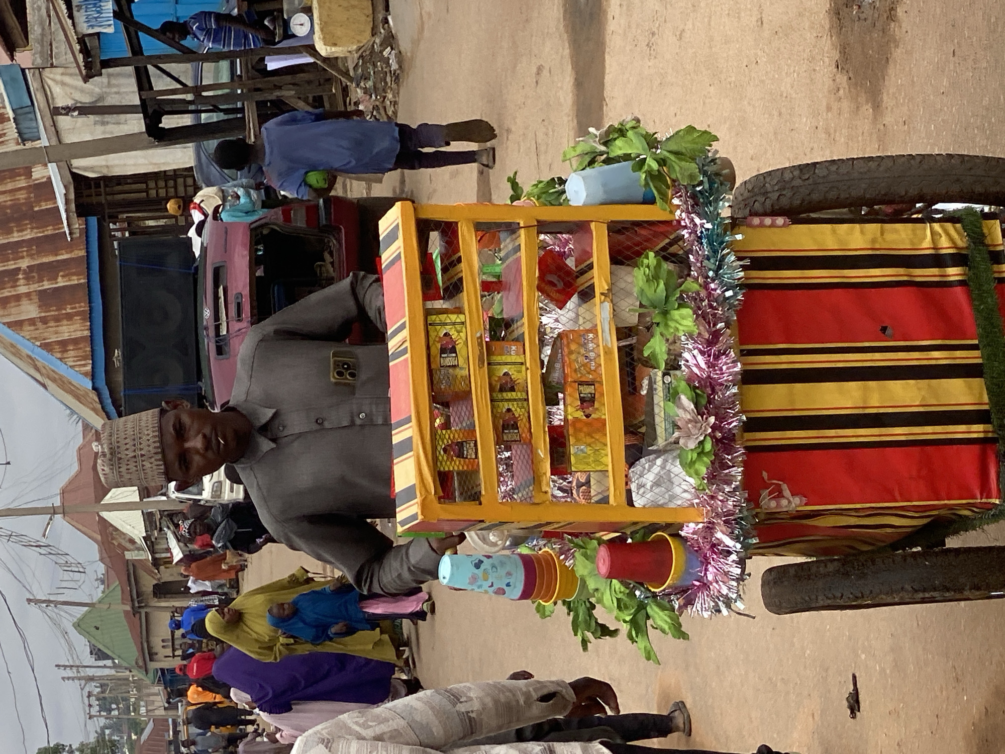 A street vendor in Rigachikun, Nigeria, stands behind a colorful cart filled with various goods. The vendor, a man wearing a traditional woven cap and a gray shirt, looks directly at the camera. The cart is decorated with red, yellow, and black stripes, and topped with greenery and festive tinsel. Stacked cups in blue and orange are visible on the left side of the cart. The background depicts a bustling street scene with people in traditional clothing, buildings with corrugated metal roofs, and power lines overhead. A red vehicle with its trunk open is parked in the middle ground. The ground is a dusty, unpaved surface, suggesting a typical Nigerian market environment.