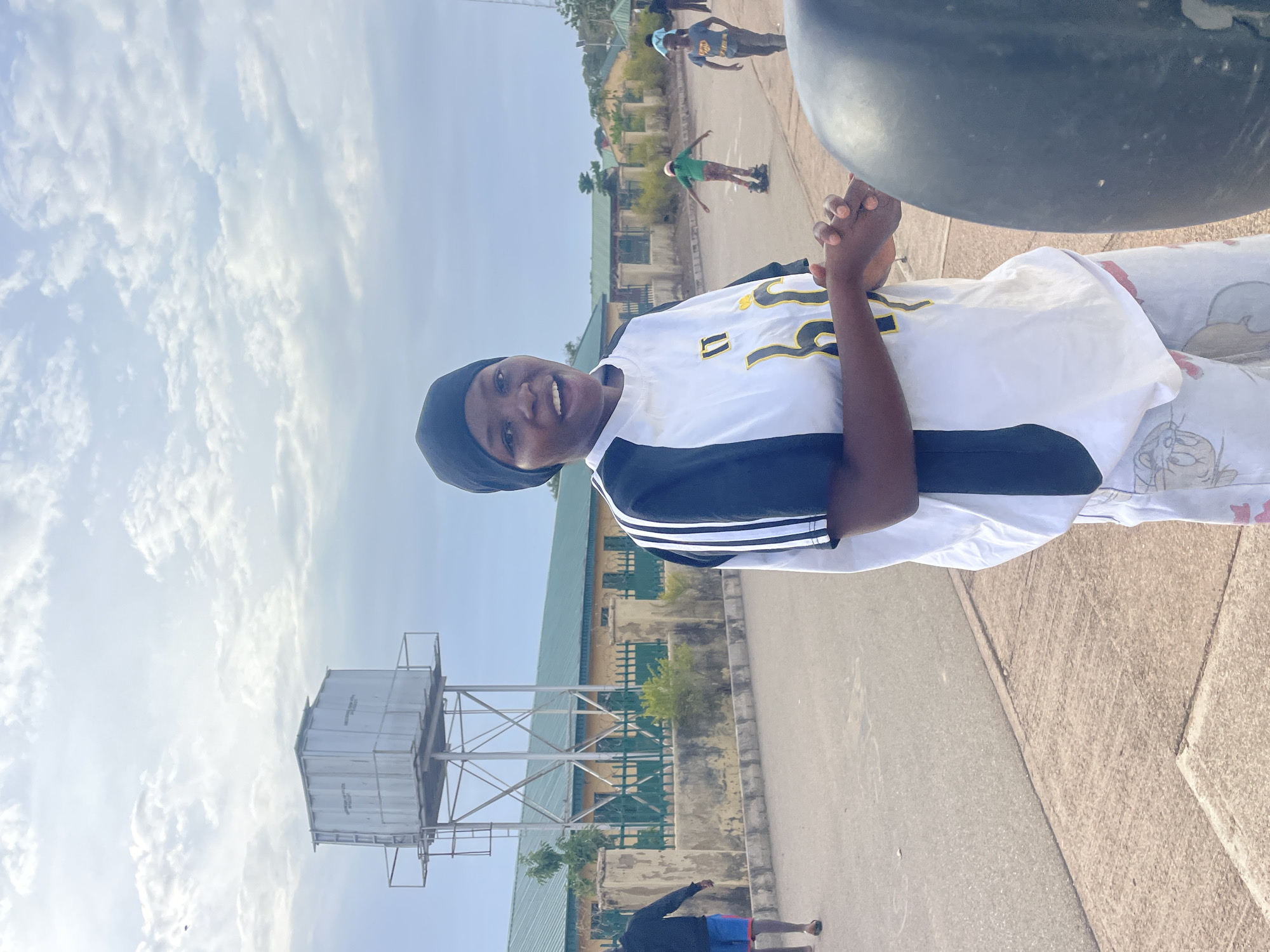 An outdoor scene in Jos, Nigeria, shows a young woman smiling at the camera in the foreground. She wears a black head covering and a white and black short-sleeved top with yellow stylized lettering, including a prominent "U," across the chest. Her hands are clasped in front of her. A large, dark, rounded object partially obstructs the upper right portion of the image, near her right side.
The setting appears to be a paved area, possibly a schoolyard or recreational ground, with a mix of asphalt and concrete surfaces. In the middle ground, a tall metal water tower with a rectangular tank stands on the left. In the background, several low-rise buildings with distinctive green roofs are visible, suggesting an institutional or educational complex.
Several other individuals are scattered in the background. One person in a green shirt and brown pants stands with arms outstretched. Another person in a blue shirt is further away on the right, and a person in a black long-sleeved top and blue shorts is partially visible in the lower left.
The image depicts a bright, daytime scene under a partly cloudy sky, indicating good weather. The main subject's cheerful smile conveys a positive and relaxed emotion. The text on the water tower is not clearly legible.
