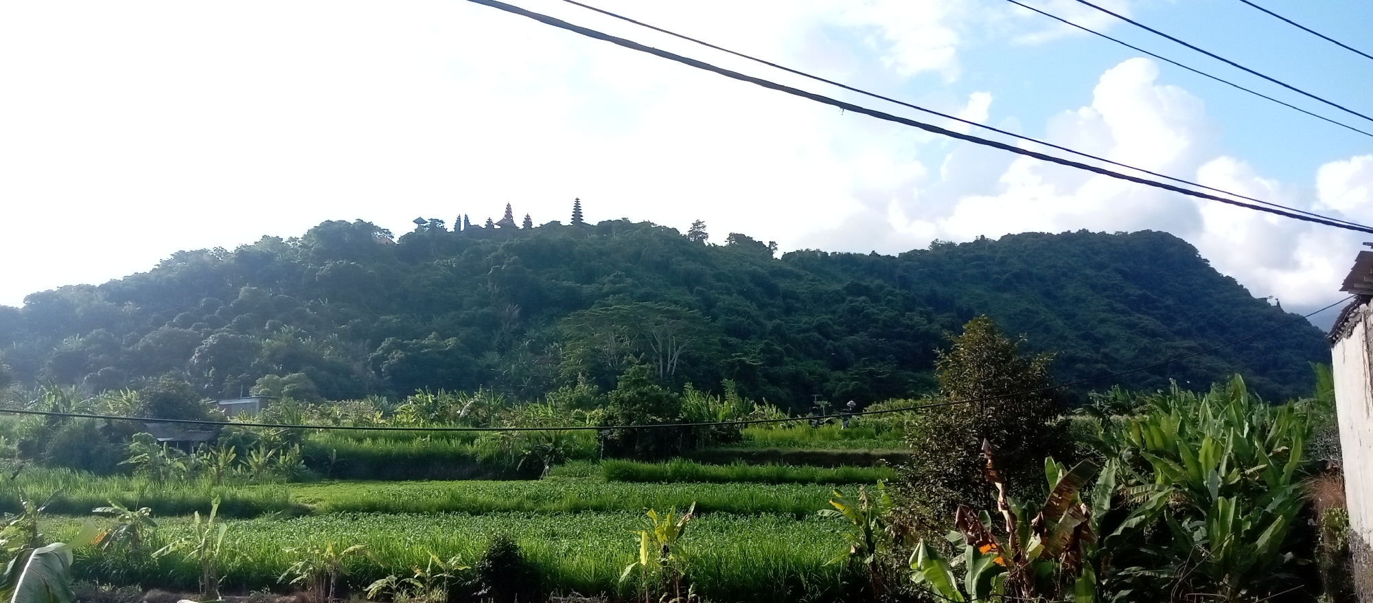 The image shows a lush green mountainous landscape with several buildings visible on the crest of the hill, suggesting it might be a religious site. In the foreground, there are terraced fields of crops, possibly rice paddies, and various types of vegetation including banana plants. The sky is partly cloudy with blue patches. Electrical wires are strung across the top of the frame. A building with a white wall is partially visible on the right side of the image. The overall scene depicts a rural or semi-rural environment in a tropical or subtropical region. Without further context or visible landmarks specific to Dawan, Indonesia, it is impossible to definitively confirm the location.