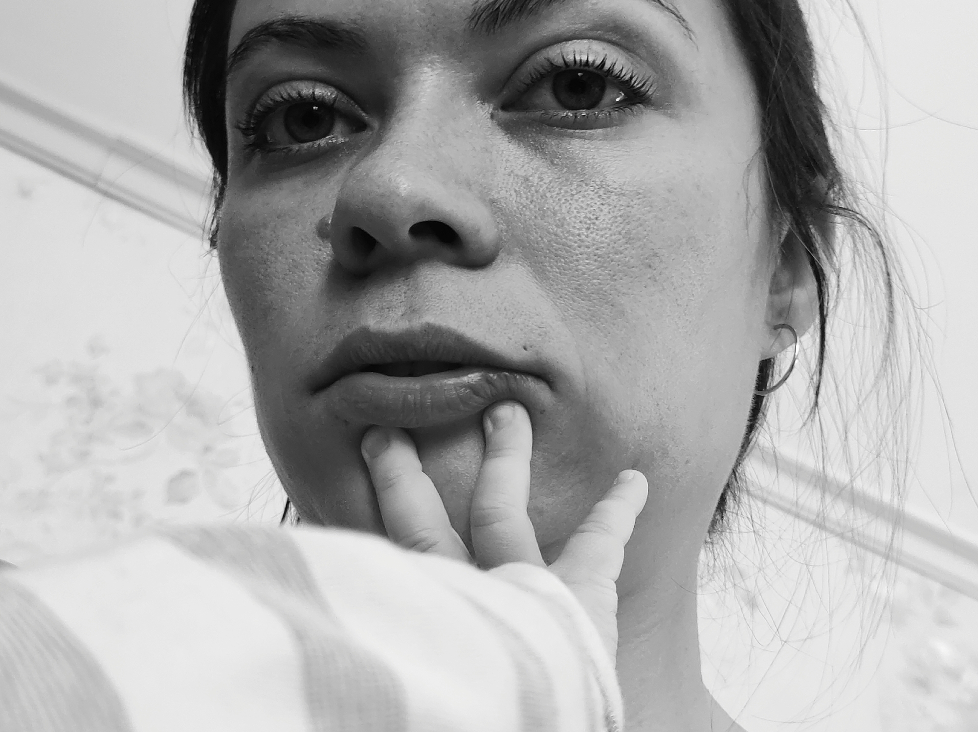 A close-up, black and white image shows a woman's face with a hand touching her lips. The woman has dark hair pulled back from her face, with a visible earring in her left ear. Her eyes are looking directly at the camera, and she has full lips. A child's hand with a striped sleeve is touching her lower lip. The background is a light-colored wall. The location is not discernible from the image.