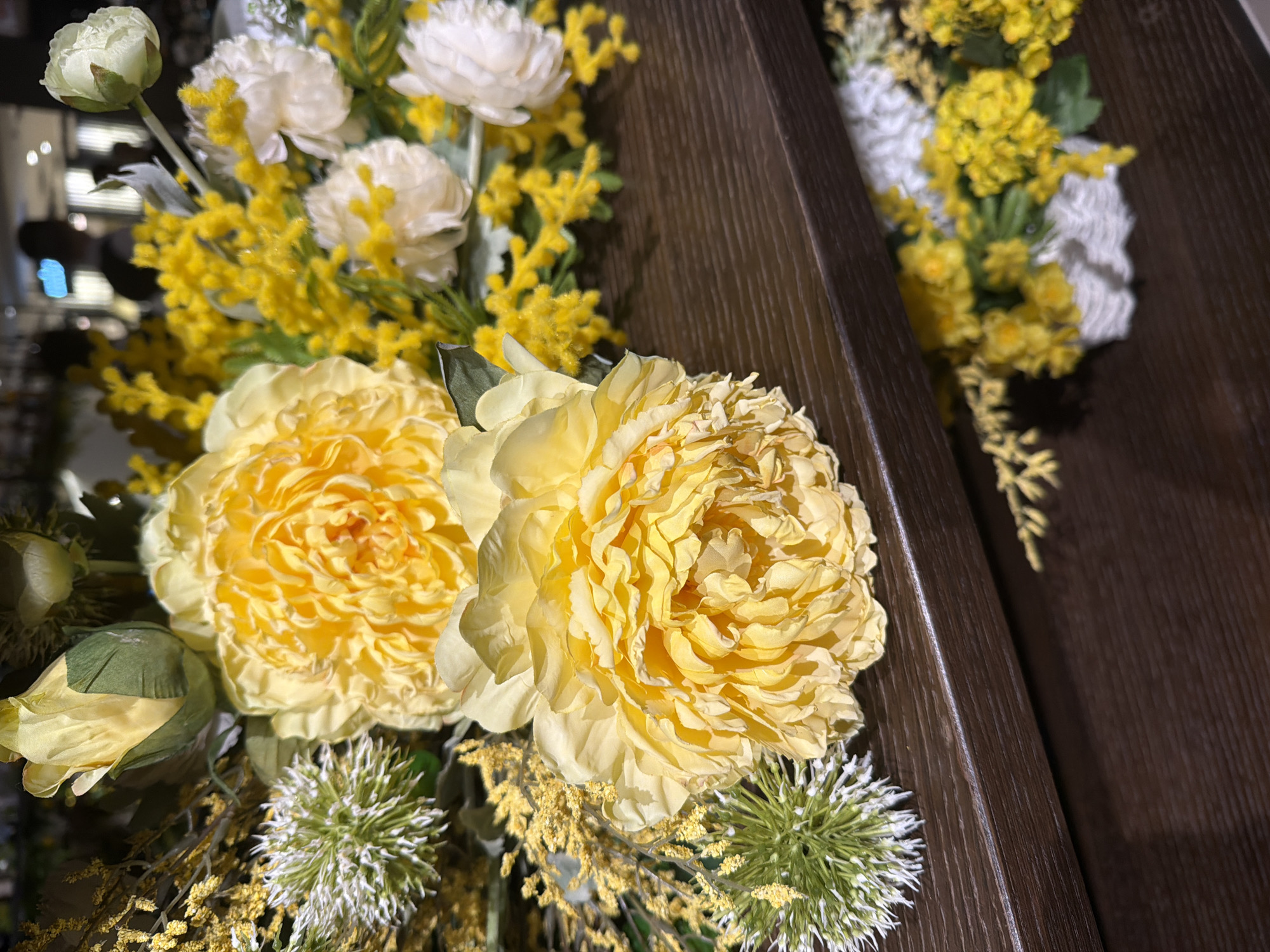 The image features a close-up view of artificial flower arrangements. Prominently displayed are two large, yellow peony-like flowers, each with densely layered petals. There are also white carnation-like flowers and various yellow and green filler flowers. Some of the filler flowers appear to be small, spiky globes. These arrangements sit next to a dark brown, wood-grained surface. The setting appears to be indoors with a dim light source in the background. There is no indication in the image that the location is Moscow, Russia.