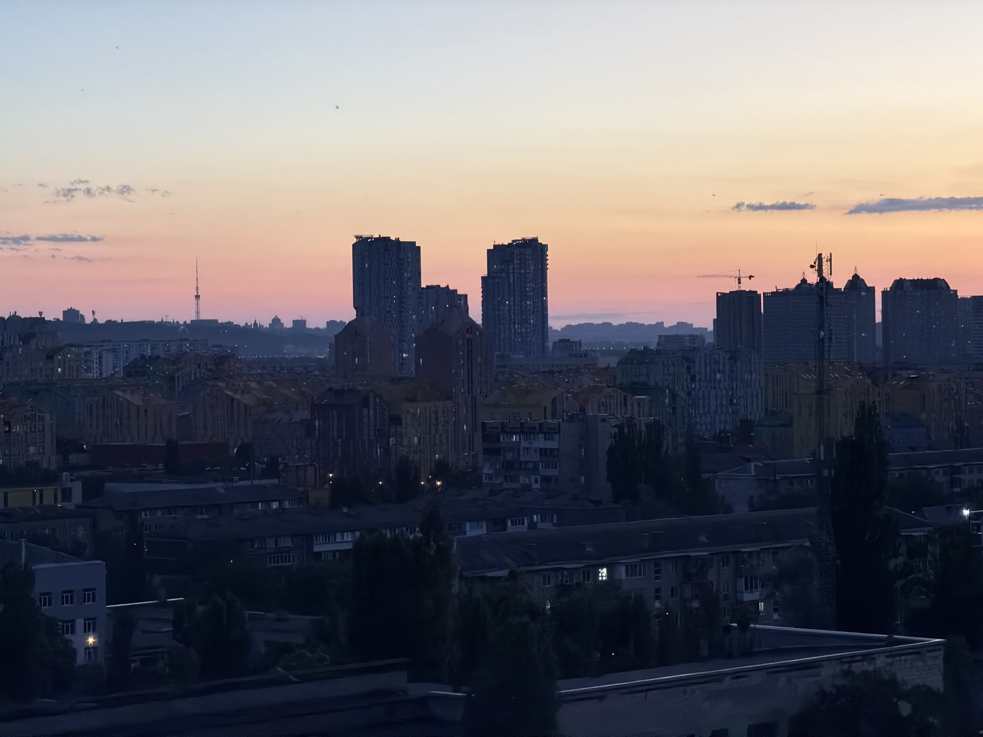 The image depicts a panoramic view of the city of Kyiv, Ukraine, at dawn or dusk, with the sky exhibiting a gradient of pastel colors from light blue to pink and orange. The cityscape is dominated by a dense arrangement of buildings, ranging from modern high-rise apartments and office buildings to older, lower-rise residential structures. In the middle ground, several tall, slender residential buildings with visible windows are clustered together. Further into the distance, a prominent radio tower with a lattice structure stands against the horizon, flanked by more urban development. The foreground shows a darker, more immediate view of residential buildings, some with illuminated windows, suggesting human presence. Trees and greenery are interspersed throughout the urban landscape, contributing to the overall scene. The atmosphere appears calm and serene, characteristic of the transitional light of twilight.