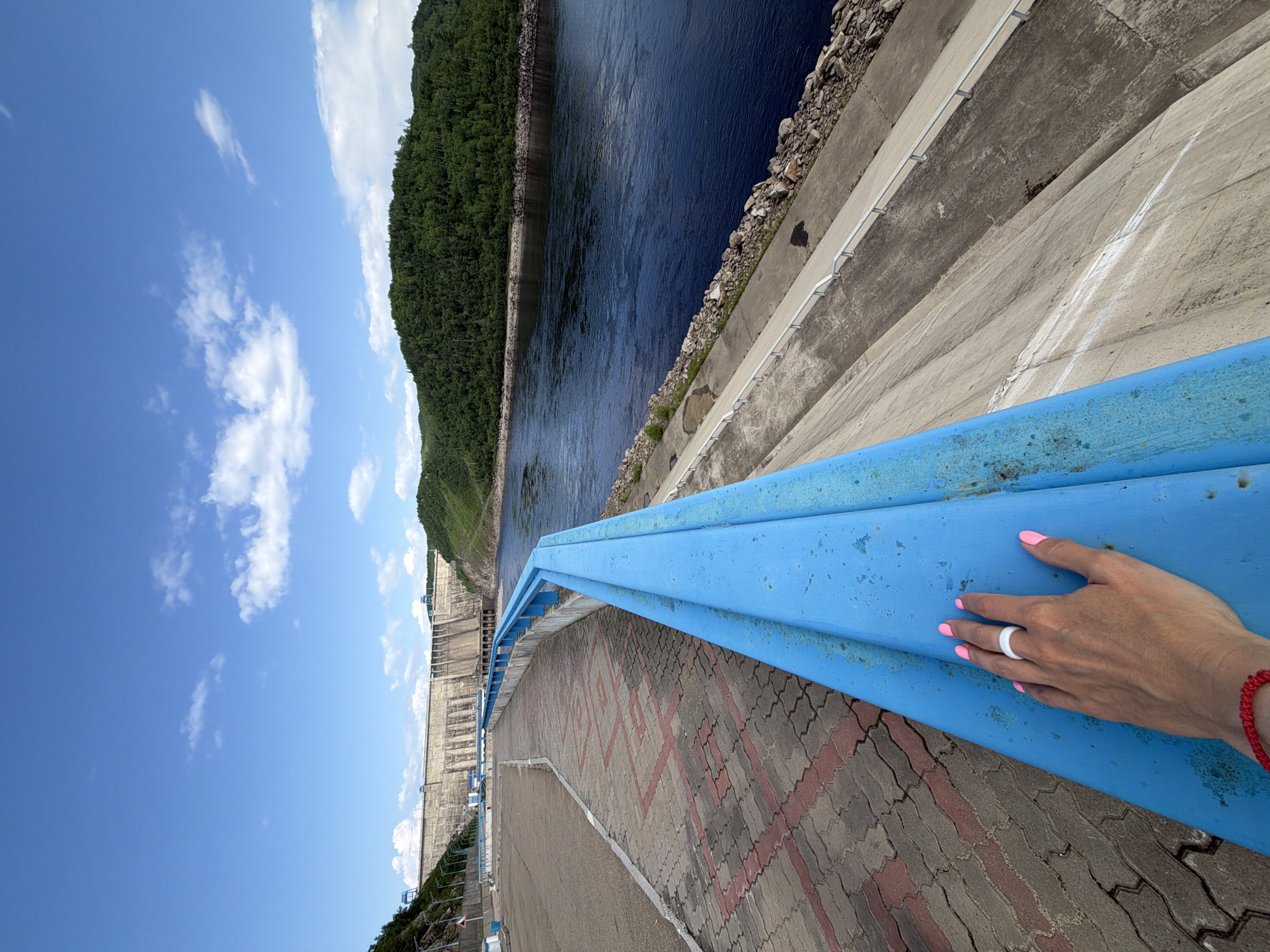 The image captures a wide view of the Zeyskaya GES (Zeya Hydroelectric Station) dam in Talakan, Russia, under a bright blue sky with scattered white clouds. A person's left hand, adorned with a white ring and bright pink nail polish, rests on a weathered blue railing in the foreground. The railing runs along a concrete embankment that slopes down towards a wide, dark blue river. The embankment is lined with rocks and a paved walkway with a patterned brick surface is visible to the left, also featuring a blue railing. In the background, the massive concrete structure of the dam stretches across the horizon, with lush green, tree-covered hills rising behind it. The scene conveys a sense of scale and the power of the hydroelectric infrastructure against the natural landscape.