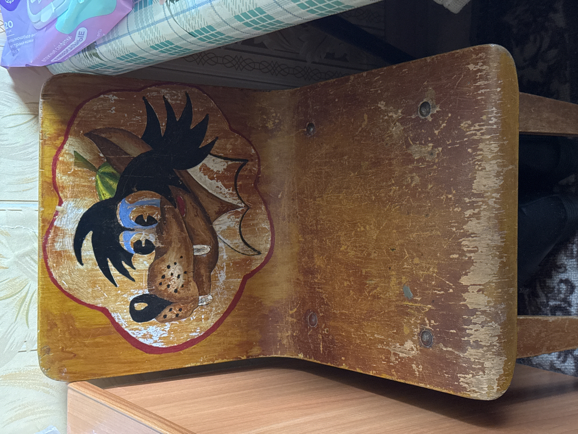 A close-up shot focuses on an old, wooden stool with a cartoonish depiction of a wolf painted on its seat. The stool appears worn, with visible scratches and chipped paint. The surrounding environment includes a tiled wall, a wooden surface, and what appears to be household items above and behind the stool. The location is specified as Chuprino, Russia.