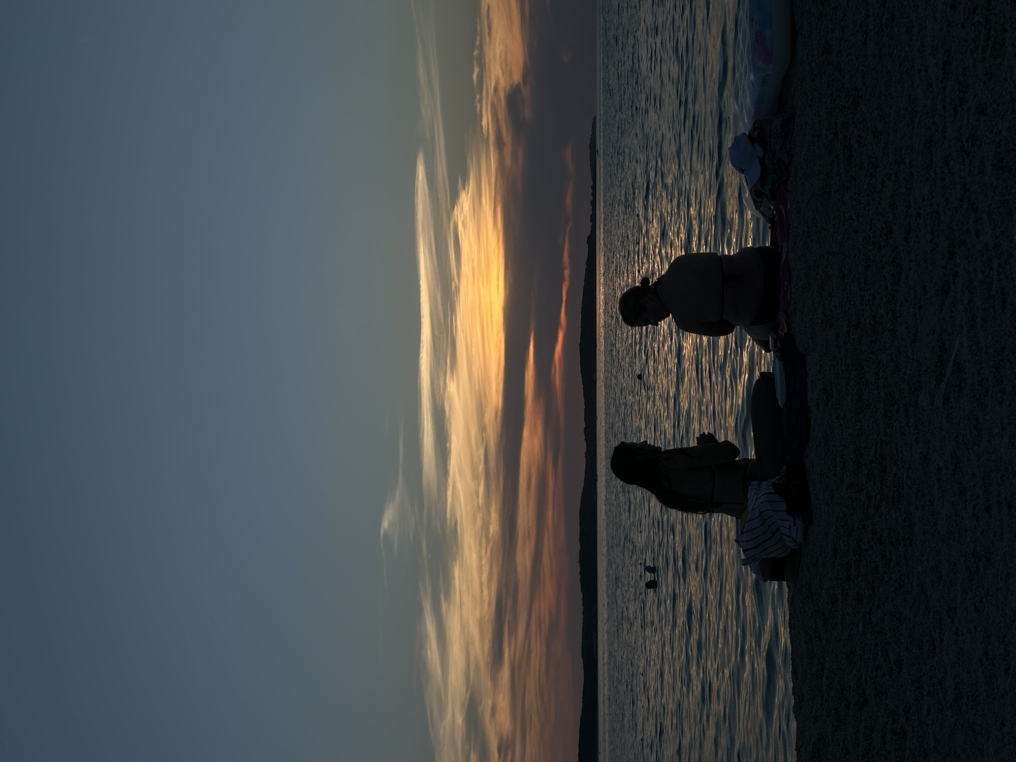 The image displays a serene sunset scene at a beach. Two individuals are silhouetted against the shimmering water. One person is kneeling, while the other appears to be sitting. In the background, a vibrant sunset paints the sky with streaks of orange, yellow, and blue, suggesting it is late evening. The water is calm with gentle ripples reflecting the sky's colors. A dark shoreline, possibly covered in sand or pebbles, occupies the right side of the frame. There are no visible objects or text in the image. The overall mood is peaceful and tranquil, capturing a moment of quiet reflection by the sea.