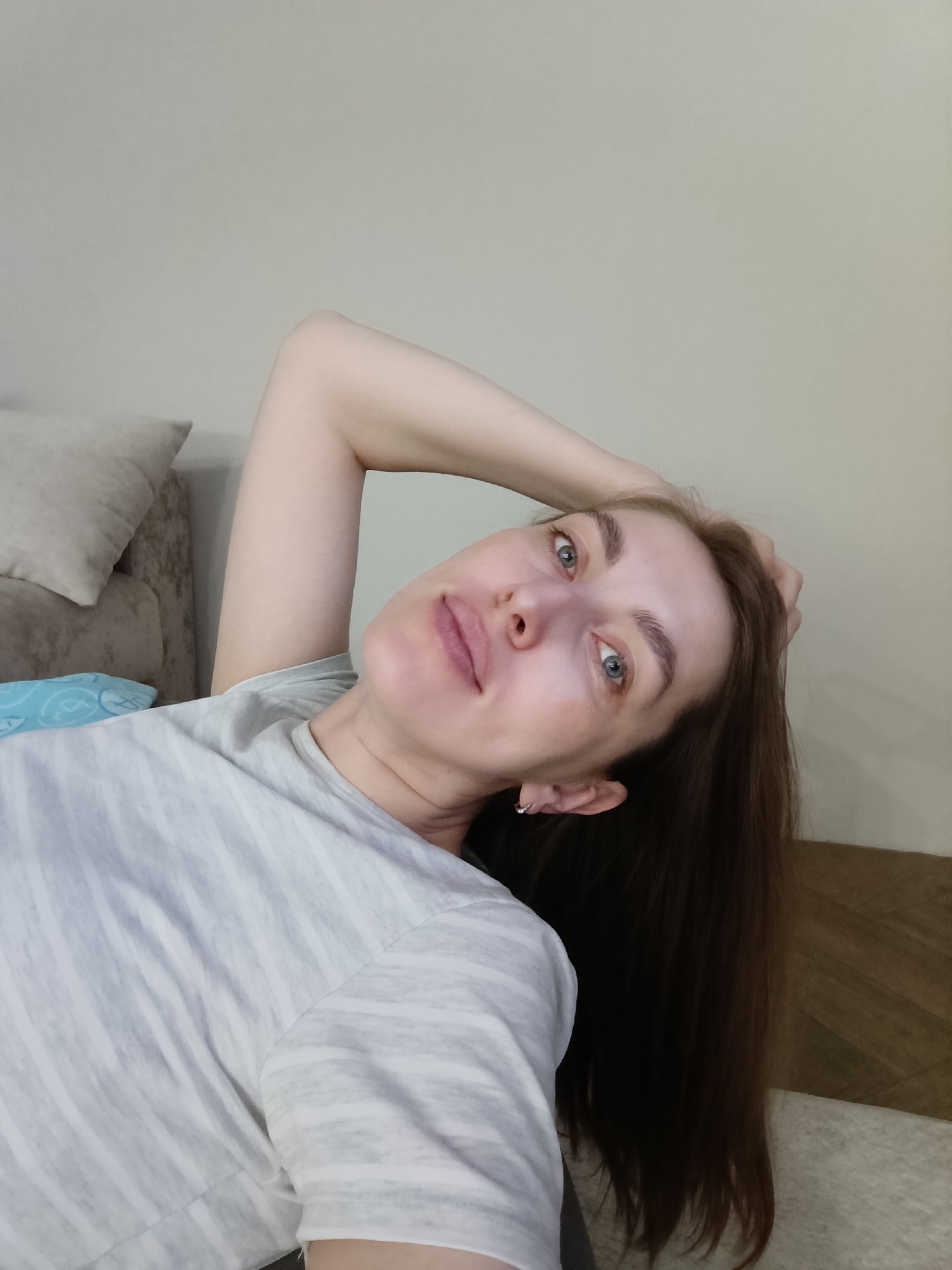 A woman with fair skin, light blue eyes, and brown hair is lying down. She is propped up with a pillow on a sofa or bed, her right arm is raised and bent behind her head. She is wearing a grey and white striped t-shirt. There is a beige wall behind her and a wooden-look surface to the right. The location is not discernible from the image alone, and the statement that the city is Chaikovsky, Russia is unsubstantiated by visual evidence.