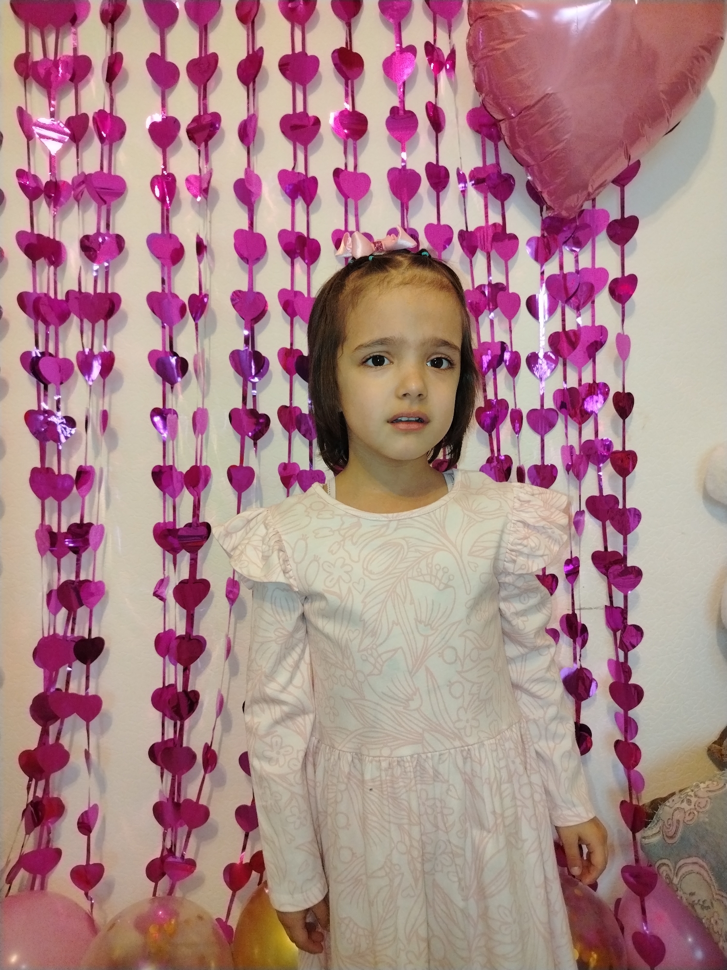 A young girl stands in front of a backdrop of pink heart-shaped decorations and a pink heart-shaped balloon. She is wearing a long-sleeved light pink dress with a floral pattern. The location is indicated as Noviy, Russia; however, this is likely an error as 'Noviy' on its own is not a city. The scene is likely indoors, possibly for a celebration, given the decorations and the girl's attire. There is additional decor and balloons partially visible at the bottom of the frame. The girl's expression is neutral.