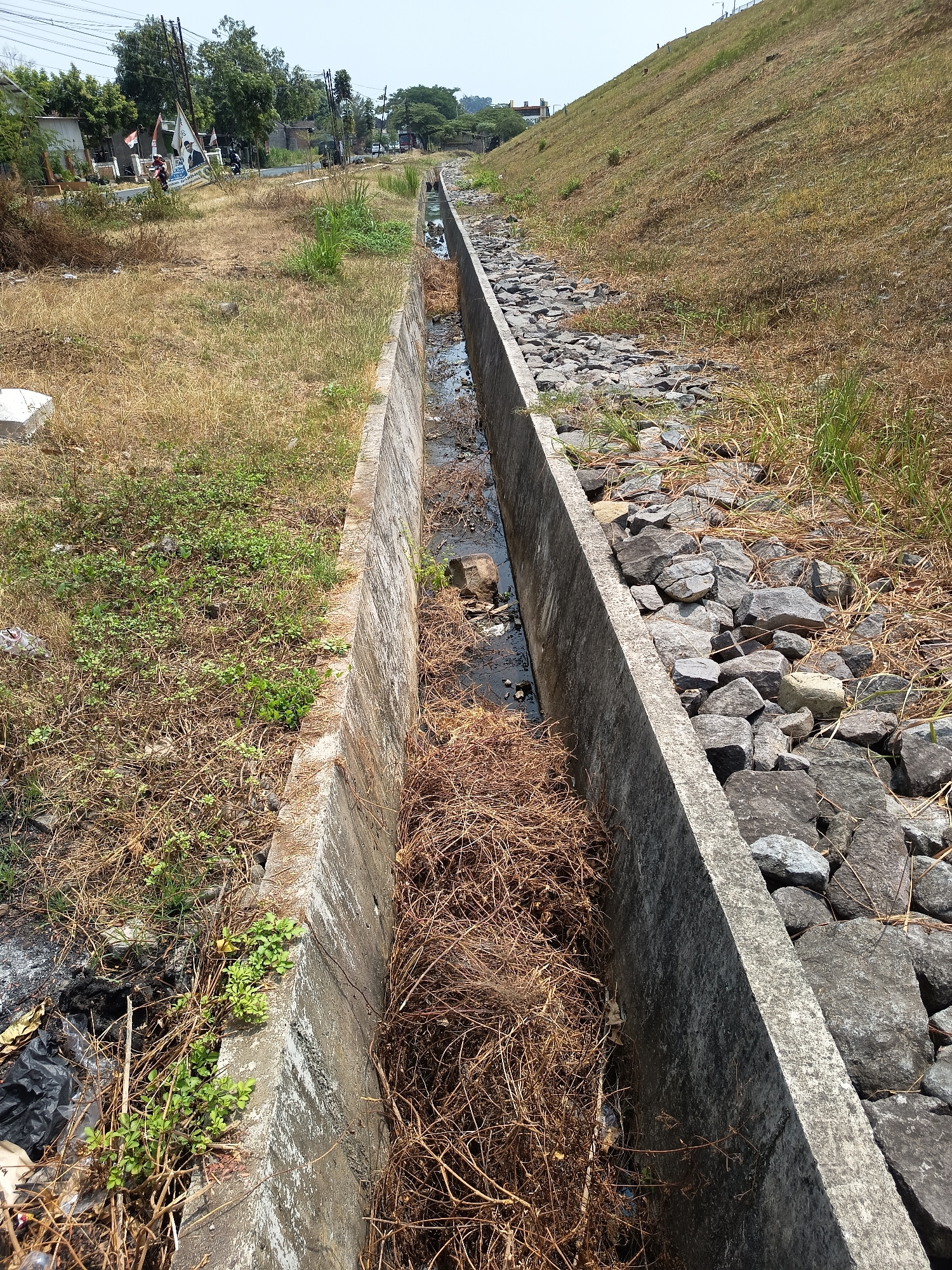 The image shows a concrete drainage channel running through a dry, grassy area. The channel is partially filled with water, debris, and dead vegetation. On one side of the channel, there is a steep, grassy embankment. On the other side, there are more rocks and dry grass. In the background, there are buildings, trees, and a road with vehicles. The scene appears to be in an urban or semi-urban area of Lalung, Indonesia, given the presence of buildings and infrastructure. The overall impression is one of a neglected or undeveloped area.
