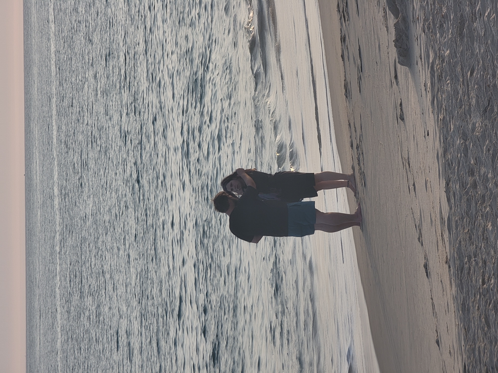 Two individuals, a male and a female, stand on a sandy beach in Quarteira, Portugal, facing the ocean. The male, wearing a black t-shirt and blue shorts, has his arms around the female, who is wearing a dark dress. The waves gently lap the shore, and the sky is a pale pink, suggesting either sunrise or sunset. The beach is mostly empty, with only footprints visible in the sand. The overall atmosphere is serene and peaceful, capturing a moment of quiet intimacy between the couple.
