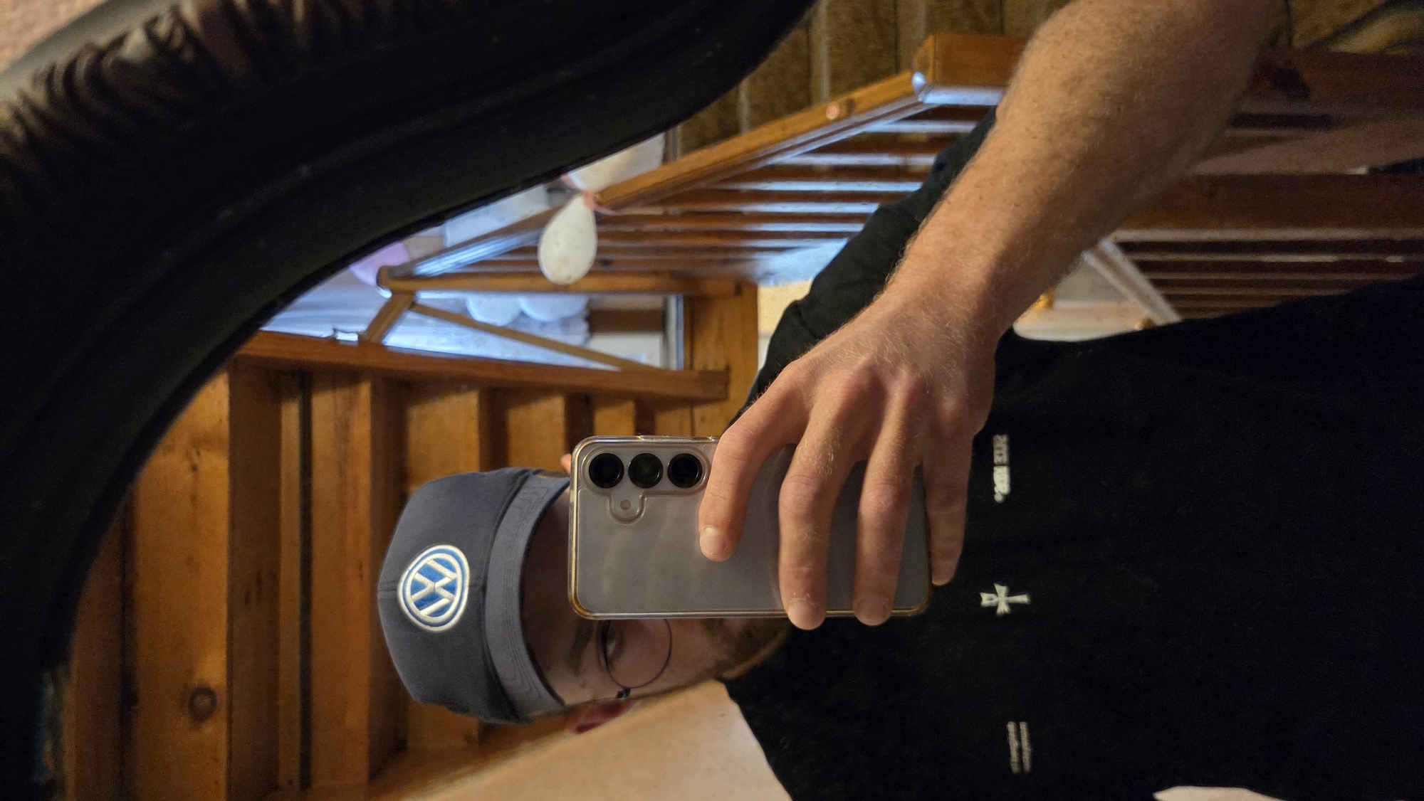 A person is taking a mirror selfie in a home, likely a hallway or stairwell, as indicated by the wooden staircase in the background. The person is wearing a dark blue baseball cap with the Volkswagen logo, glasses, and a black t-shirt with a white symbol and text. They are holding a smartphone in their right hand to capture the reflection. The lighting suggests an indoor environment.